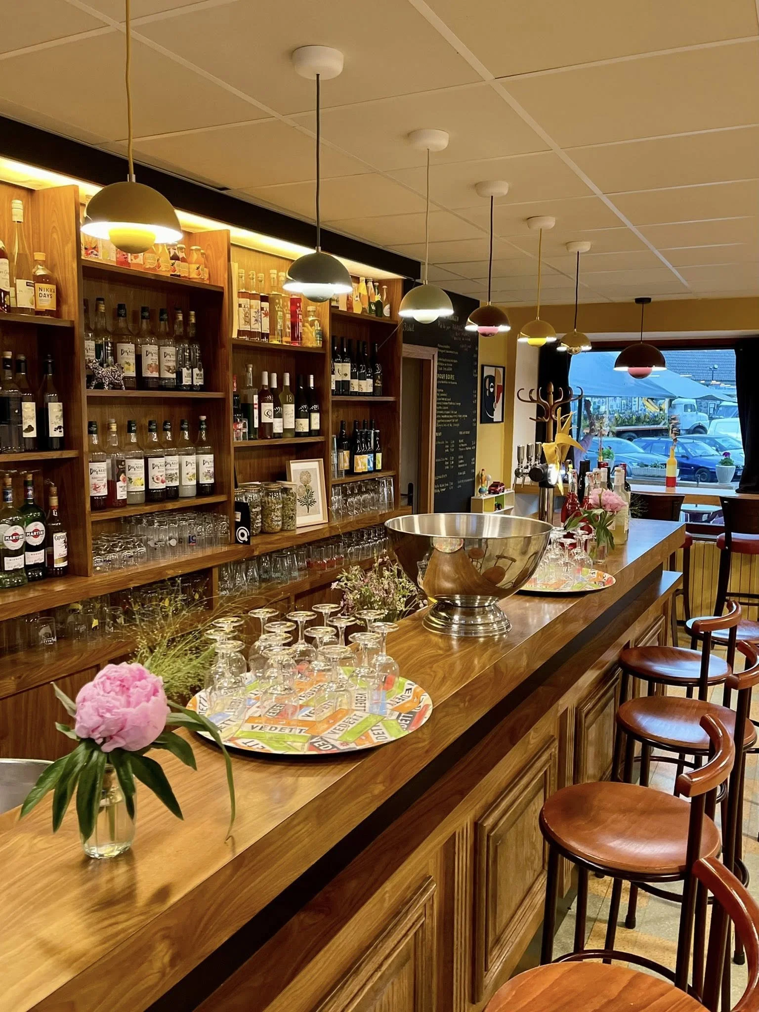 Un bar en intérieur avec une longue comptoir en bois, décoré de fleurs, verres et une grande bol en métal. En arrière-plan, des étagères en bois remplies de bouteilles de différentes boissons alcoolisées et verres. Lampes suspendues au plafond. Fenêt
