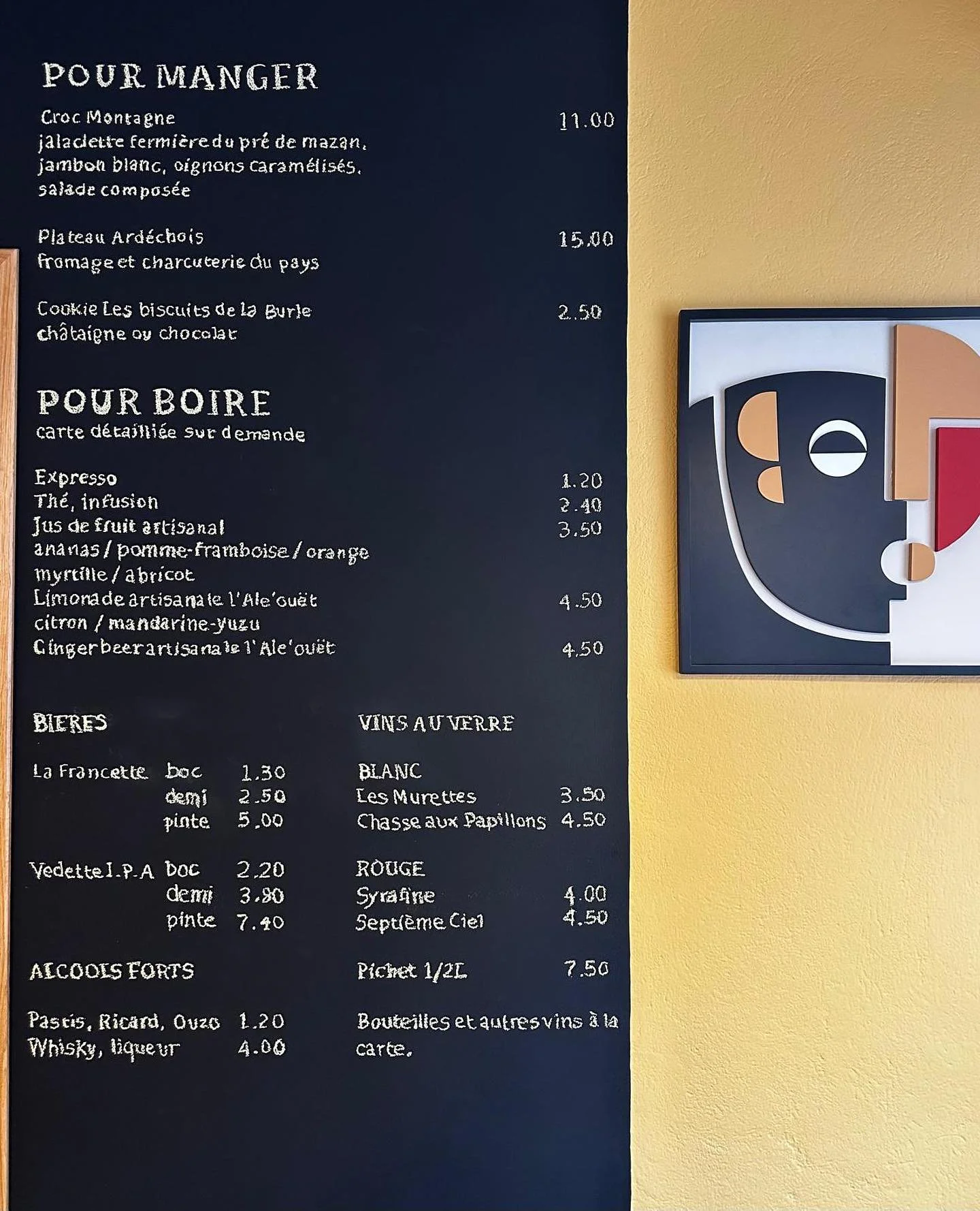 Menu en français avec options de plats, boissons, vins et alcools, écrit sur un tableau noir, avec un tableau de décoration moderne jaune à côté.