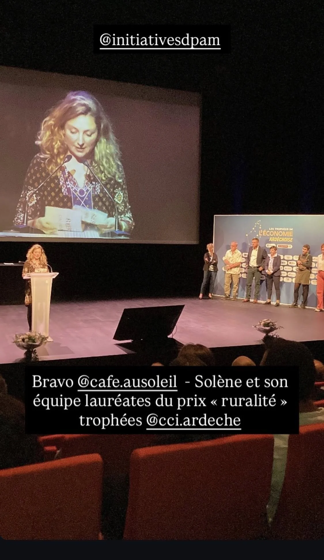 Une femme parle à une cérémonie sur scène, avec un grand écran derrière elle montrant sa silhouette. D'autres personnes sont alignées sur le côté droit de la scène. La scène est décorée avec un panneau indiquant "Les Trophées de l'Économie Ardèche". 