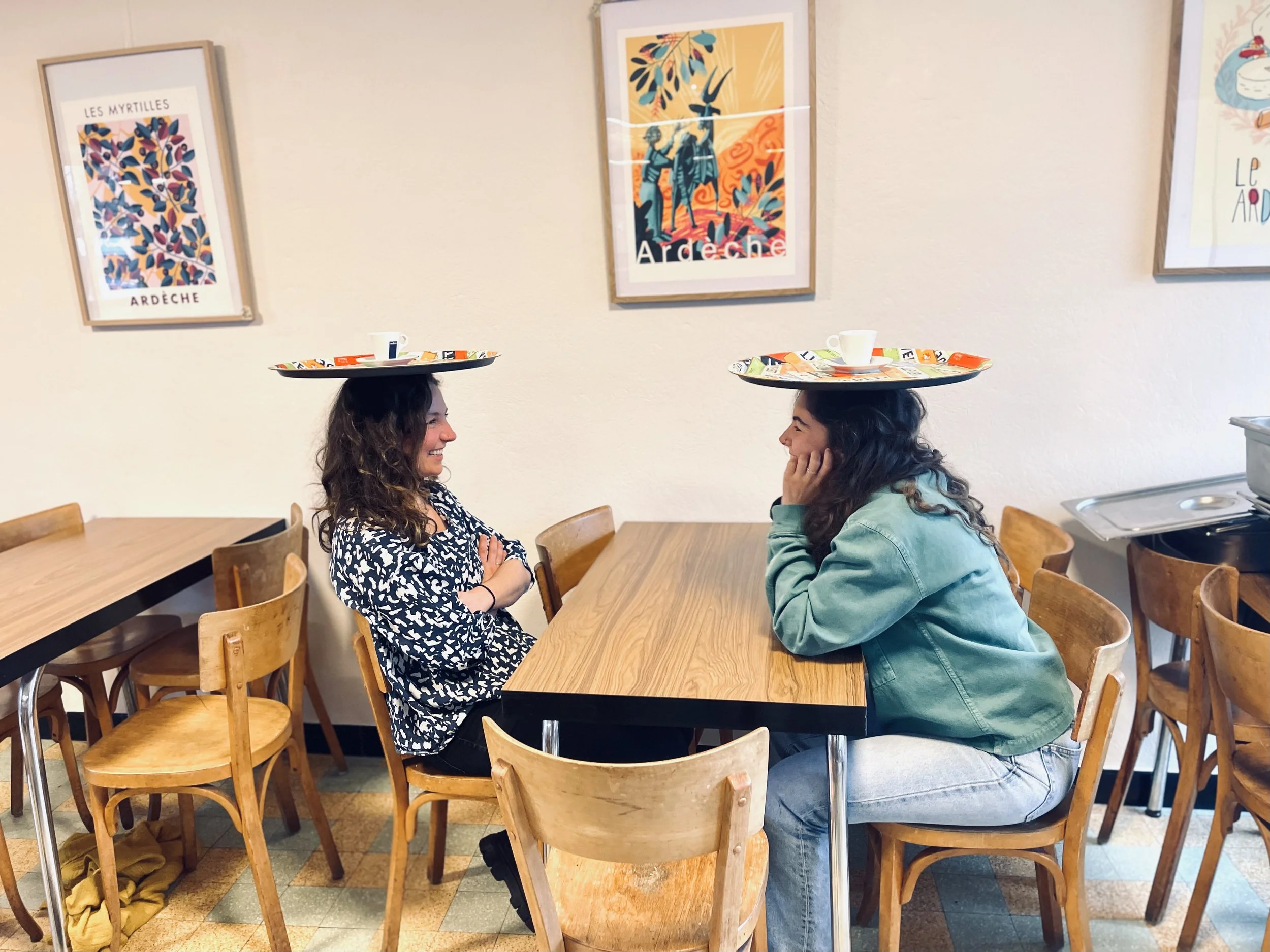 Deux femmes avec des plateaux sur la tête, assises à une table de café et se regardant mutuellement, dans un intérieur décoré avec des affiches colorées sur le mur.