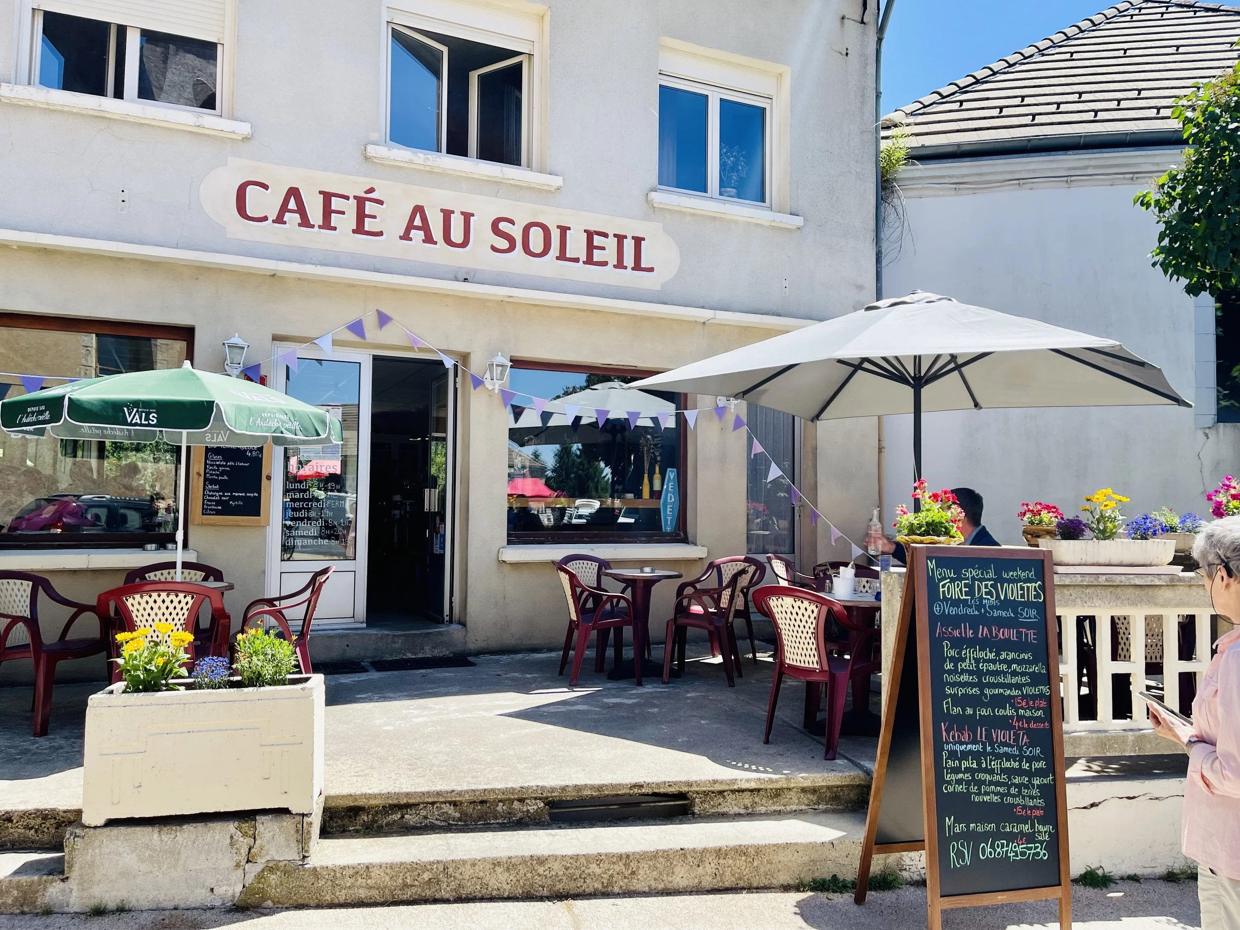 Café nommé 'Café au Soleil' avec tables extérieures, parasols, et un tableau de menu. Une femme regardant un téléphone est présente à droite.