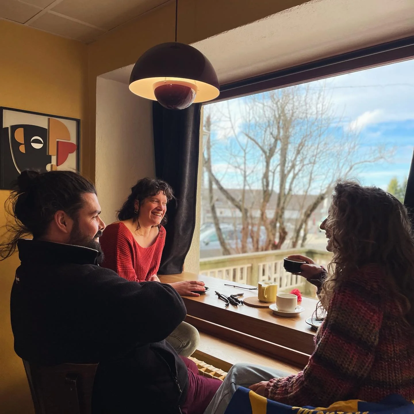 Trois personnes souriantes assises autour d'une table en bois près d'une fenêtre, en train de discuter et de boire du café ou du thé, dans une ambiance chaleureuse et lumineuse.