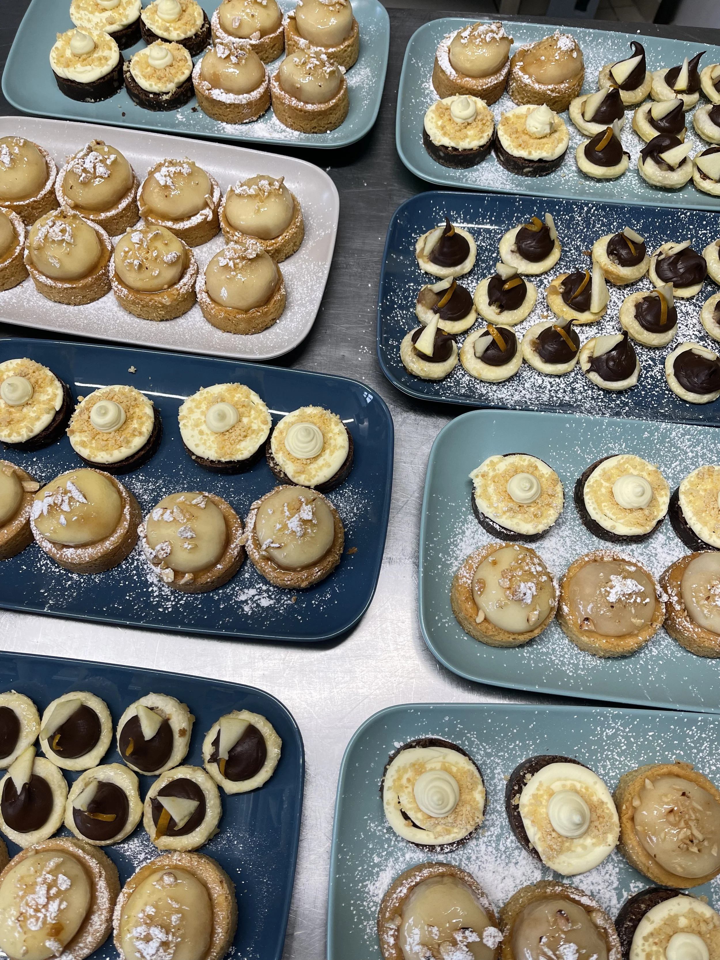 Plateaux de petits desserts variés, notamment des tartelettes au citron, des biscuits décorés avec de la crème blanche et du chocolat, tous saupoudrés de sucre glace.