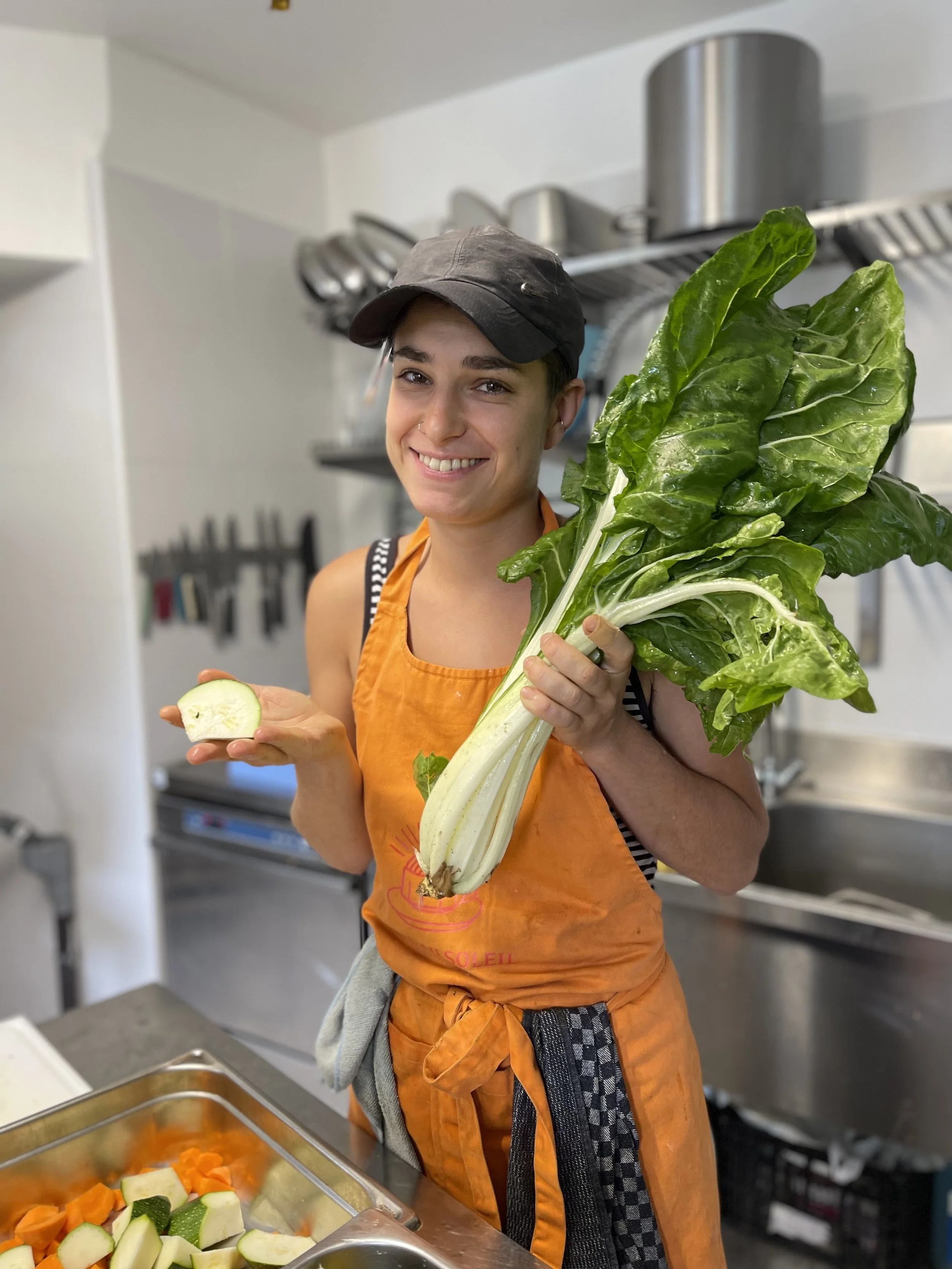 Une femme souriante tenant un bouquet de légumes verts, notamment de la bette à carde, dans une cuisine, avec une tranche de courgette dans l'autre main.