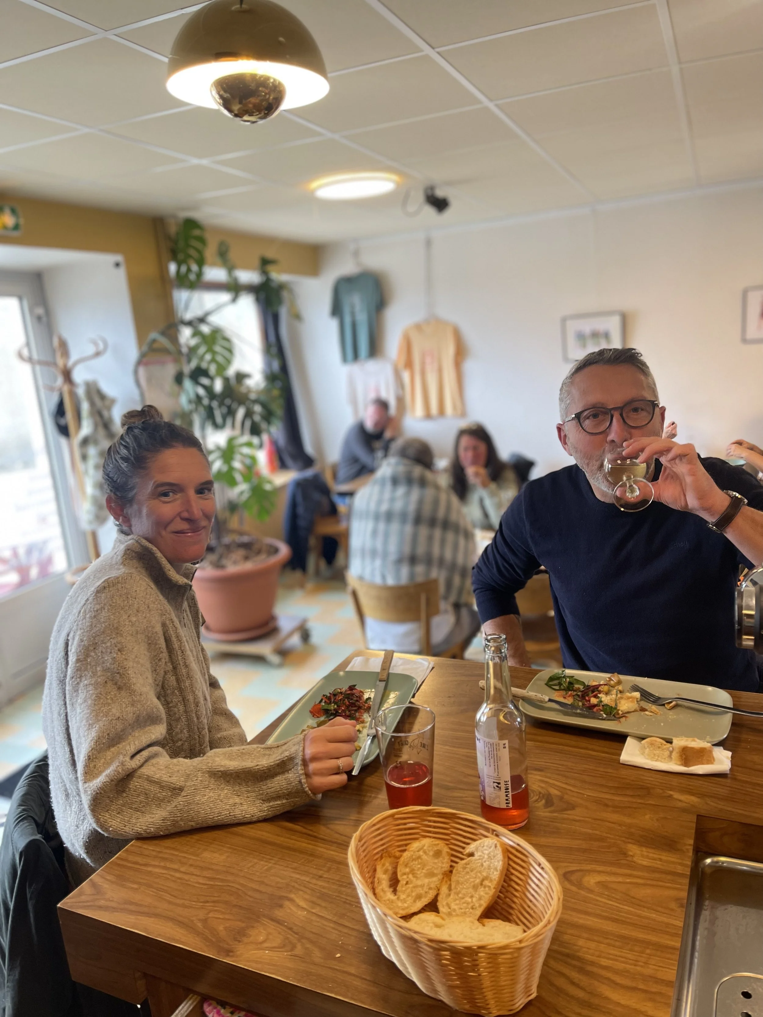 Deux personnes mangent à une table dans un restaurant, une femme souriante avec un pull beige et un homme buvant un cocktail, avec d'autres clients en arrière-plan et des vêtements accrochés au mur.