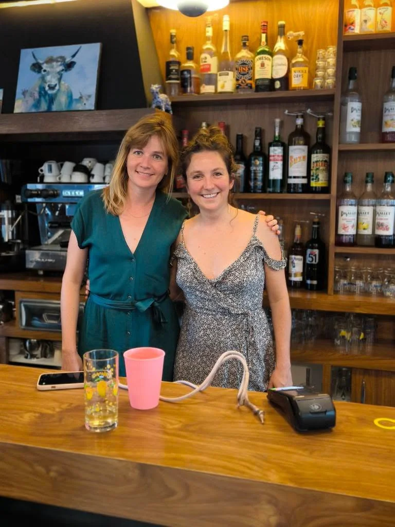 Deux femmes souriantes derrière un bar en bois, avec des bouteilles d'alcool et des verres derrière elles, dans un café ou un bar.