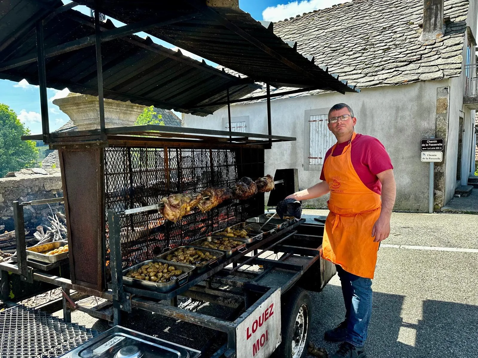 Un homme en t-shirt rouge et tablier orange cuisine avec un barbecue en plein air, préparant de la viande et des légumes, à côté d'une vieille maison en pierre.