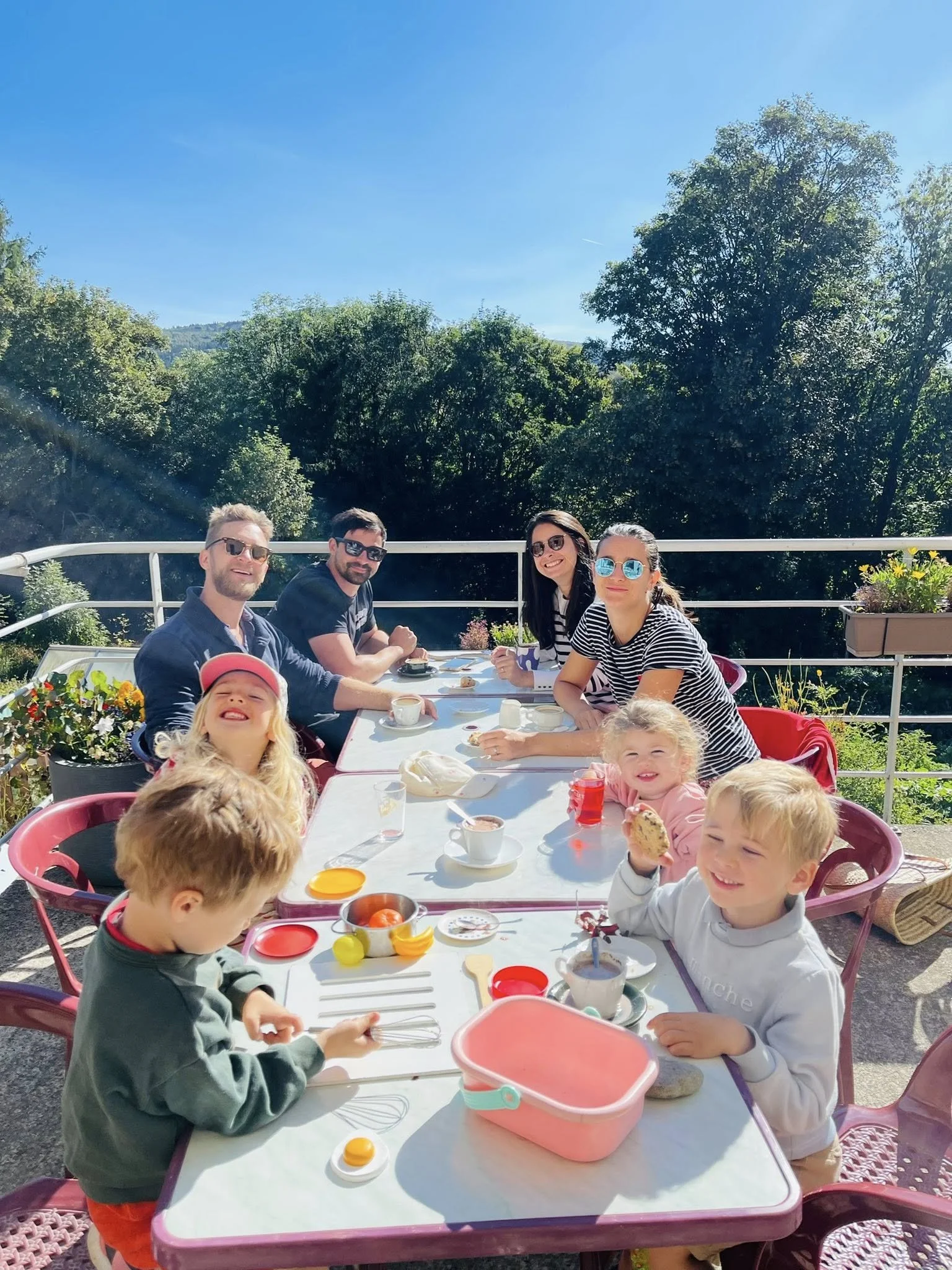 Groupe d'adultes et d'enfants assis à une table en extérieur, profitant d'une journée ensoleillée, partageant un rassemblement convivial avec nourriture et boissons, entouré de nature avec des arbres en arrière-plan.