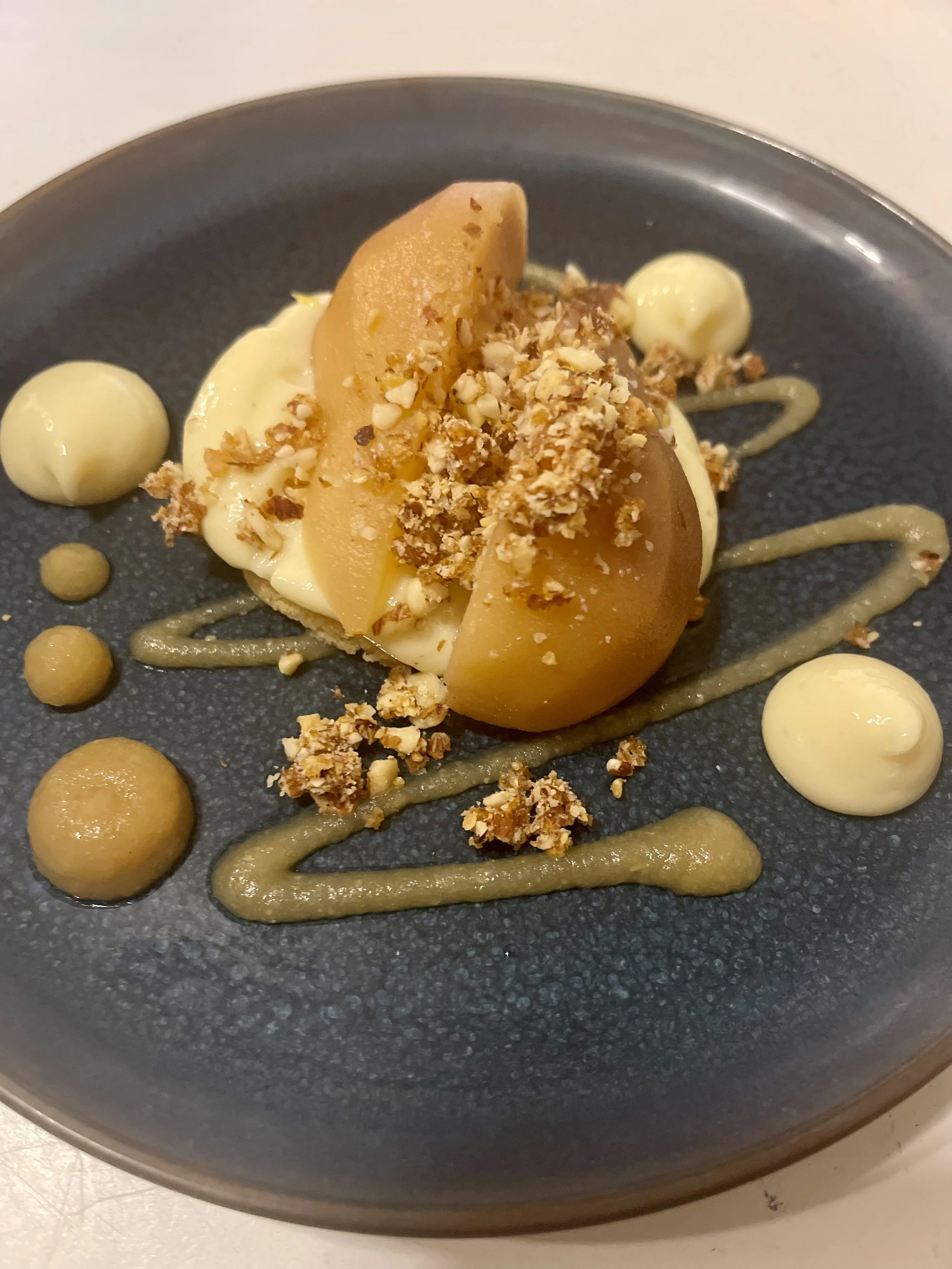 Dessert avec poire pochée, crumble, et différentes sauces et spheres crémeuses sur une assiette noire.