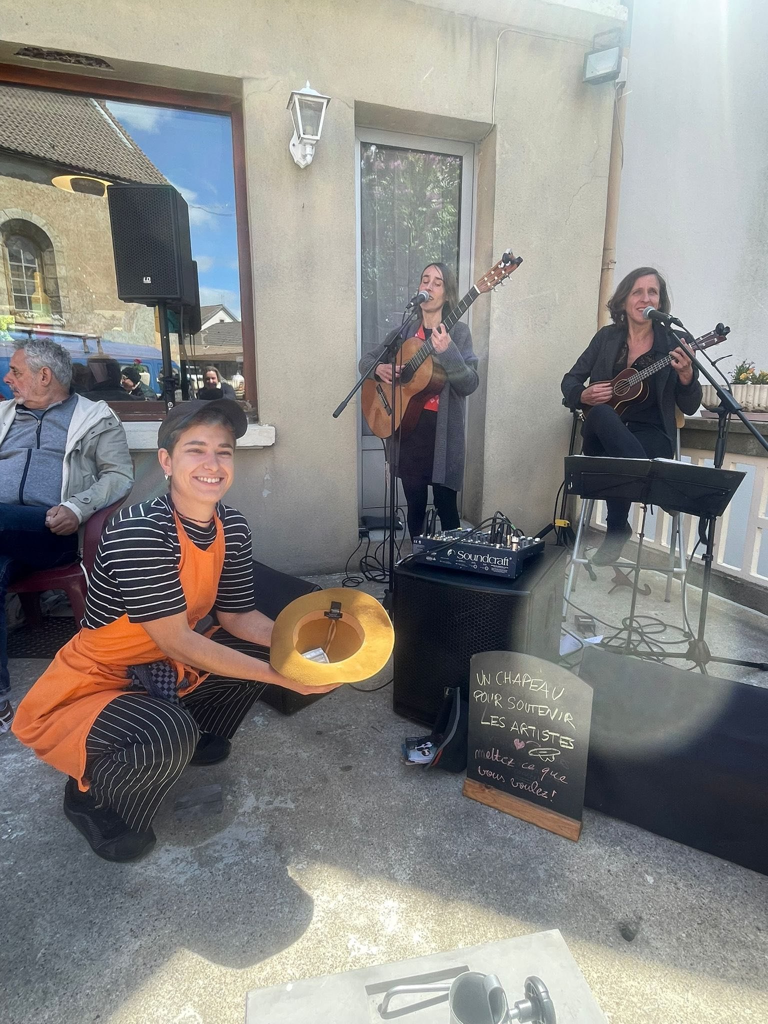 Un groupe de musiciens jouent en plein air lors d'une performance, avec une femme à gauche, souriante, tenant un chapeau doré. Deux autres femmes jouent de la guitare, une chantant dans un micro, l'autre tenant une guitare, derrière un panneau noir a