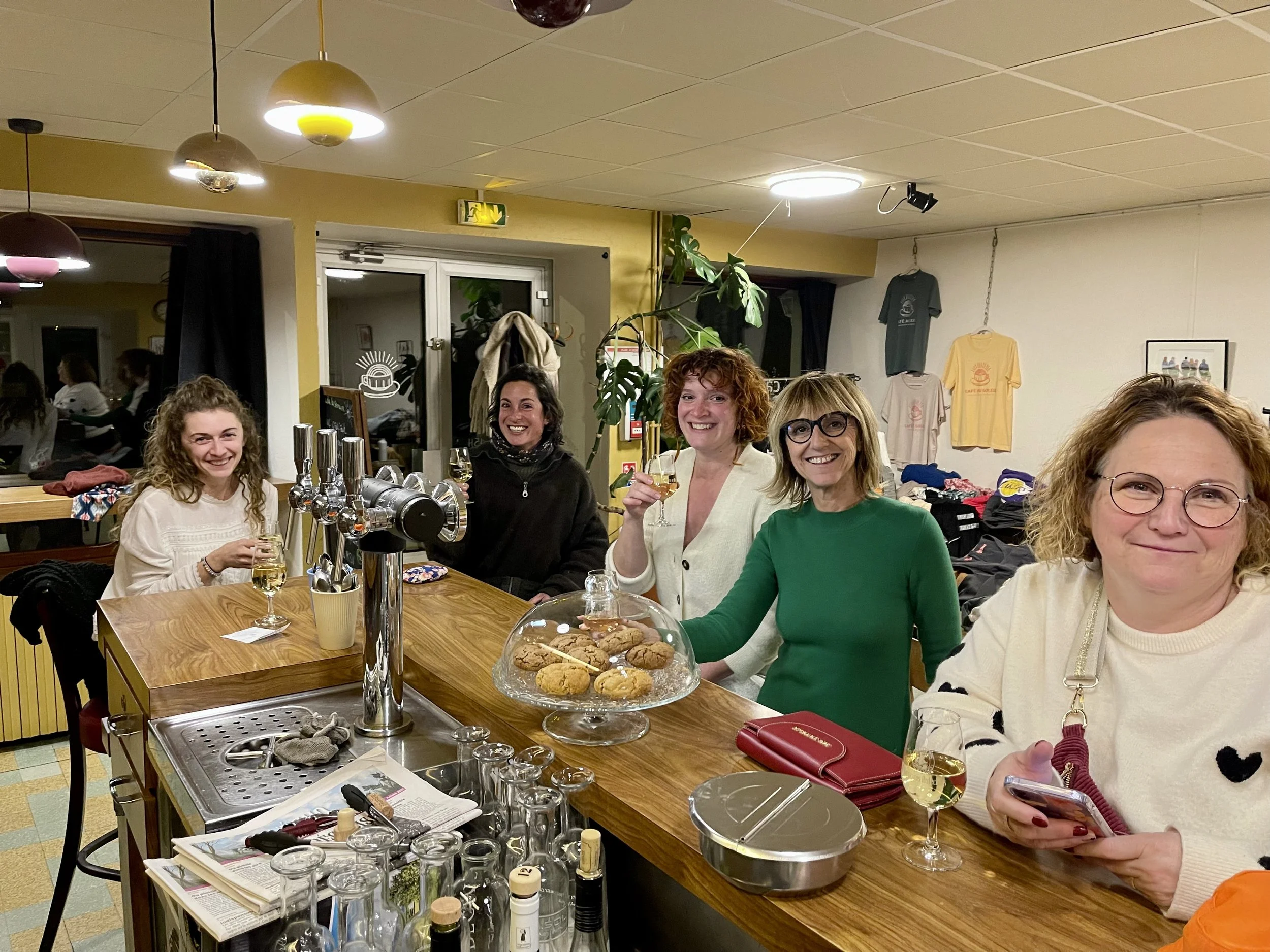 Cinq femmes souriantes et en train de boire du vin blanc autour d'un comptoir de bar dans un café ou restaurant, avec cookies en garde dans une assiette en verre.