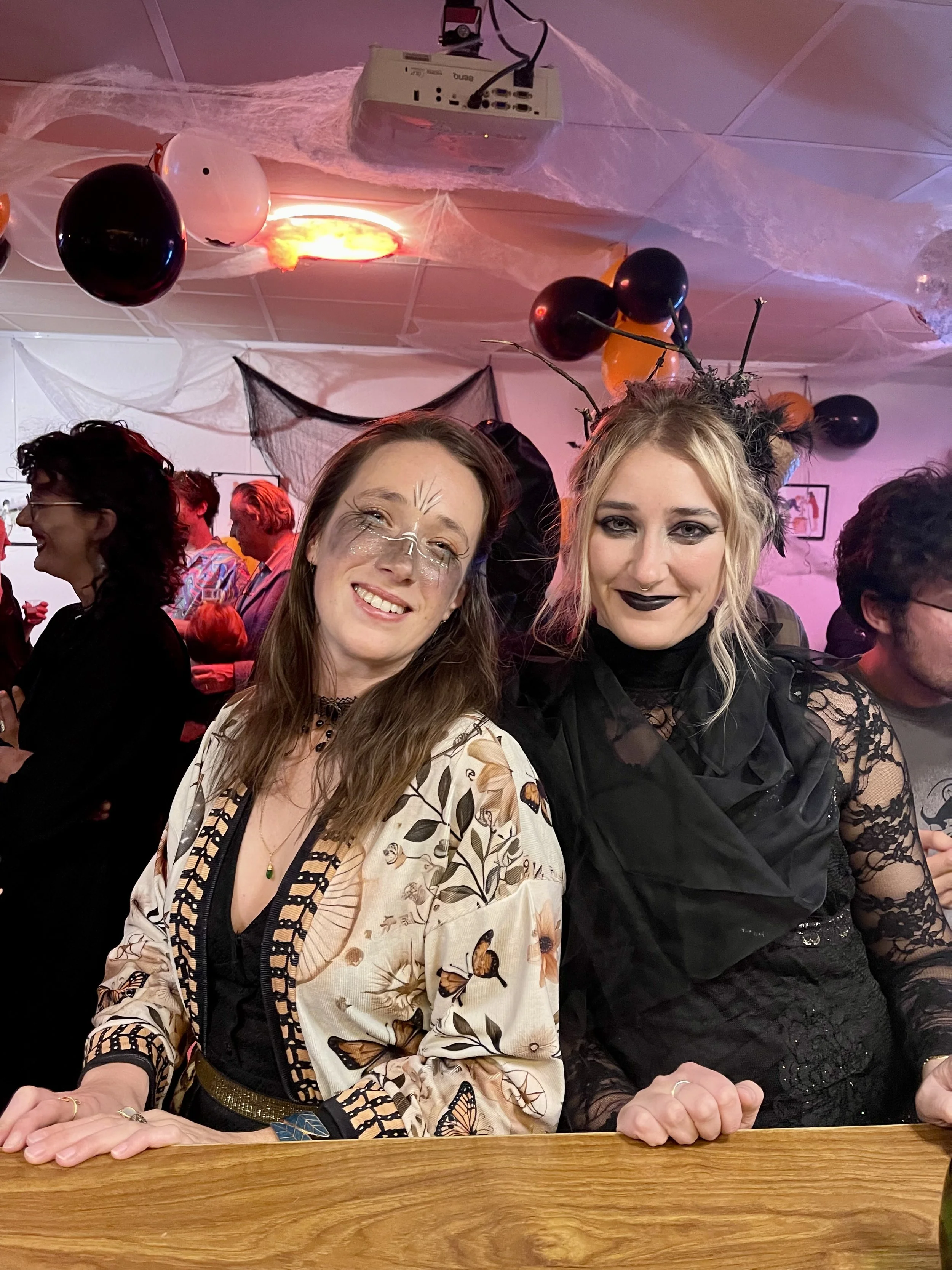 Deux femmes déguisées joyeusement lors d'une fête d'Halloween, entourées de décorations de fête et d'autres personnes en arrière-plan.