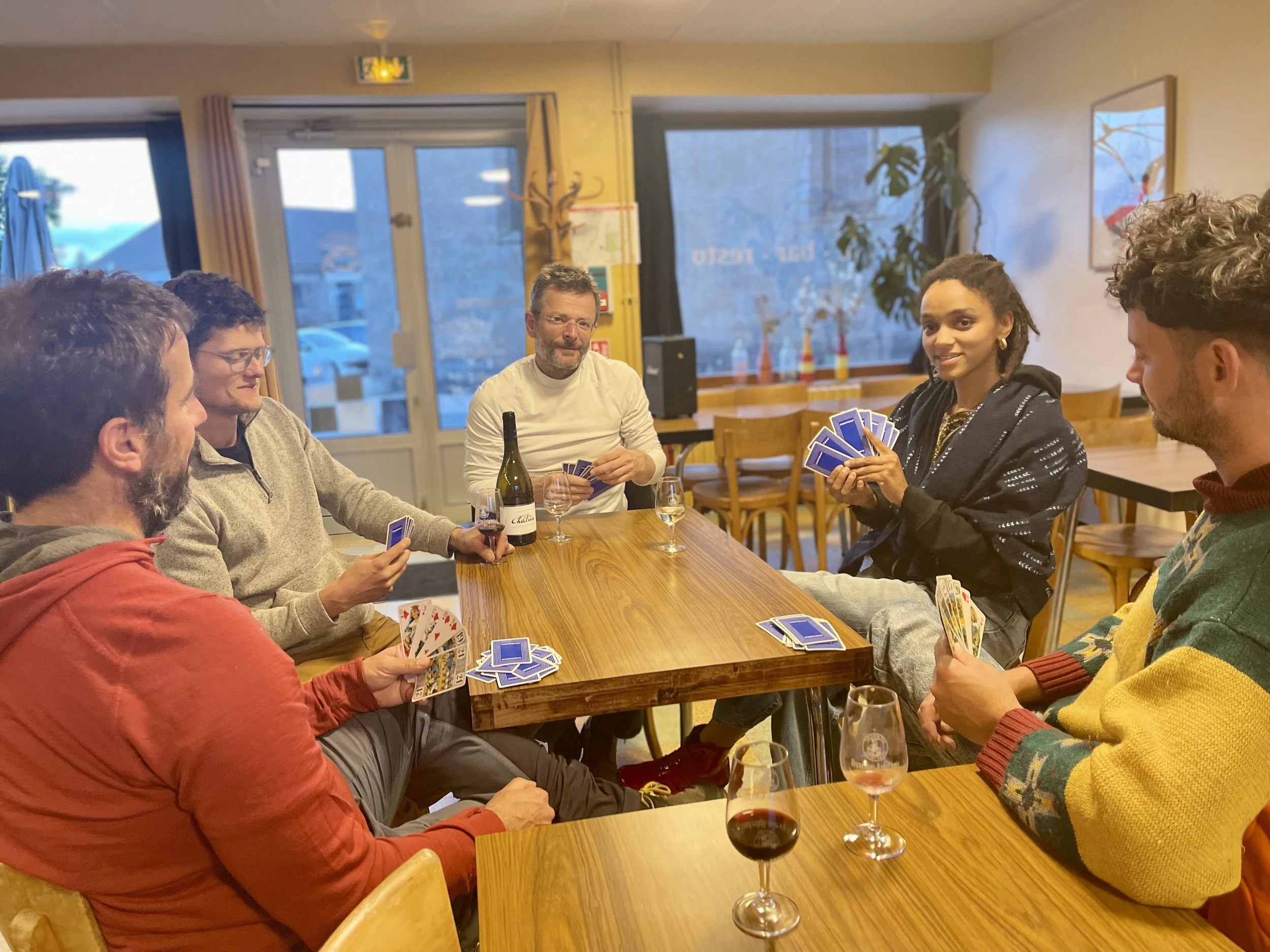 Groupe de six personnes jouant aux cartes dans un café, avec des boissons sur la table.