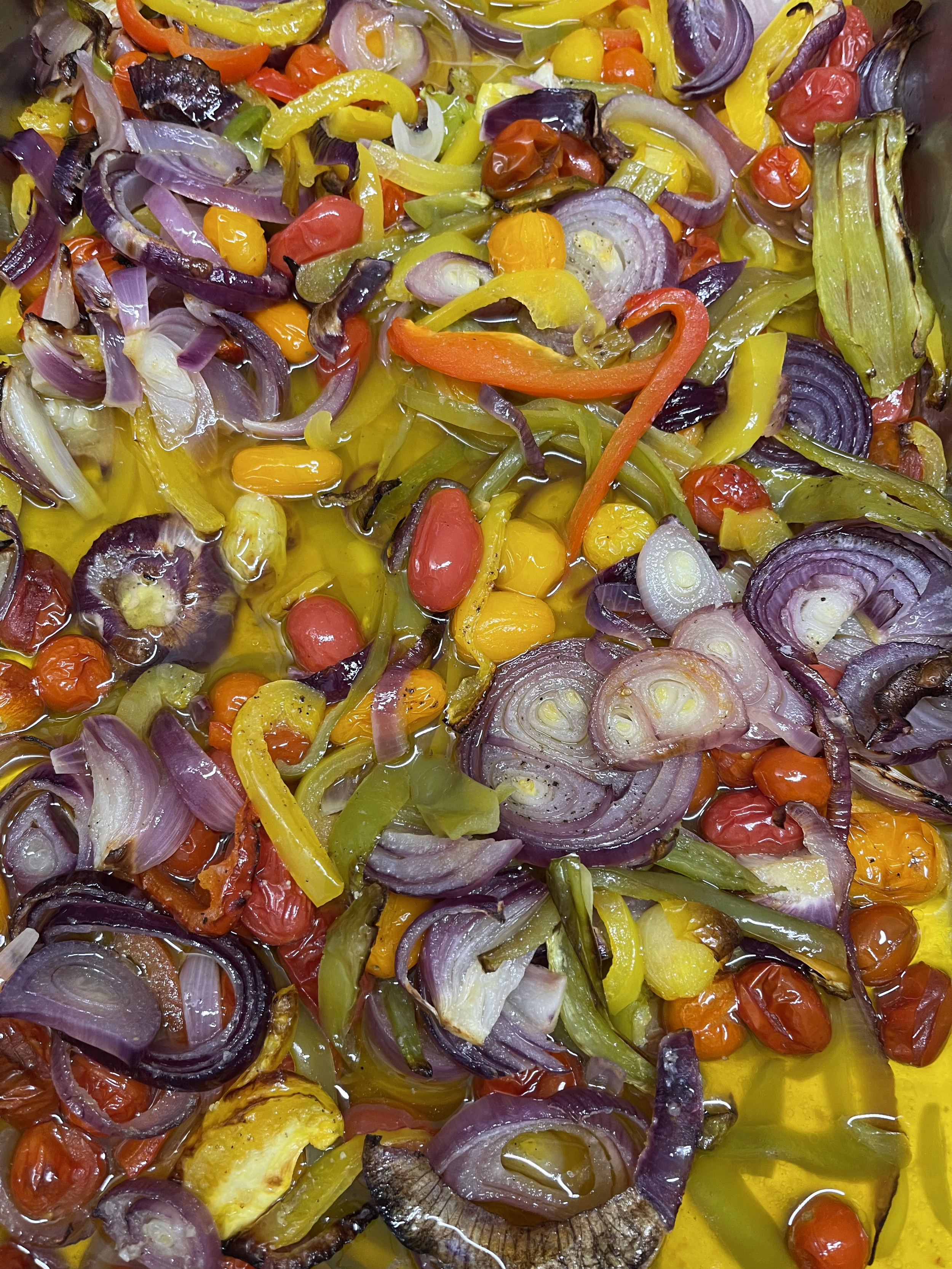 Un mélange de légumes cuits, comprenant des oignons rouges en rondelles, des poivrons jaunes et verts, des tomates cerises rouges, des petits piments rouges et jaunes, et des morceaux d'aubergine, dans de l'huile.