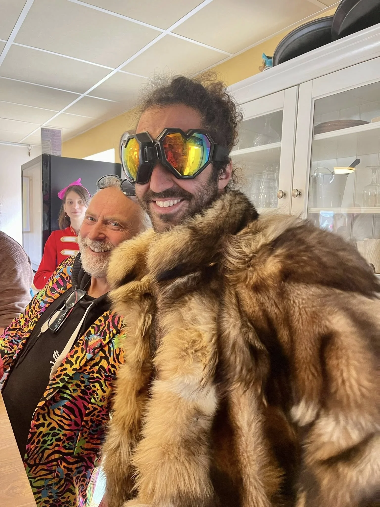 Un homme portant des lunettes de protection multicolores, un manteau en fourrure et un chien de grande taille en pelage épais accrochée à son épaule. En arrière-plan, deux autres personnes, une femme avec un hoodie à motif, et un enfant avec un bande