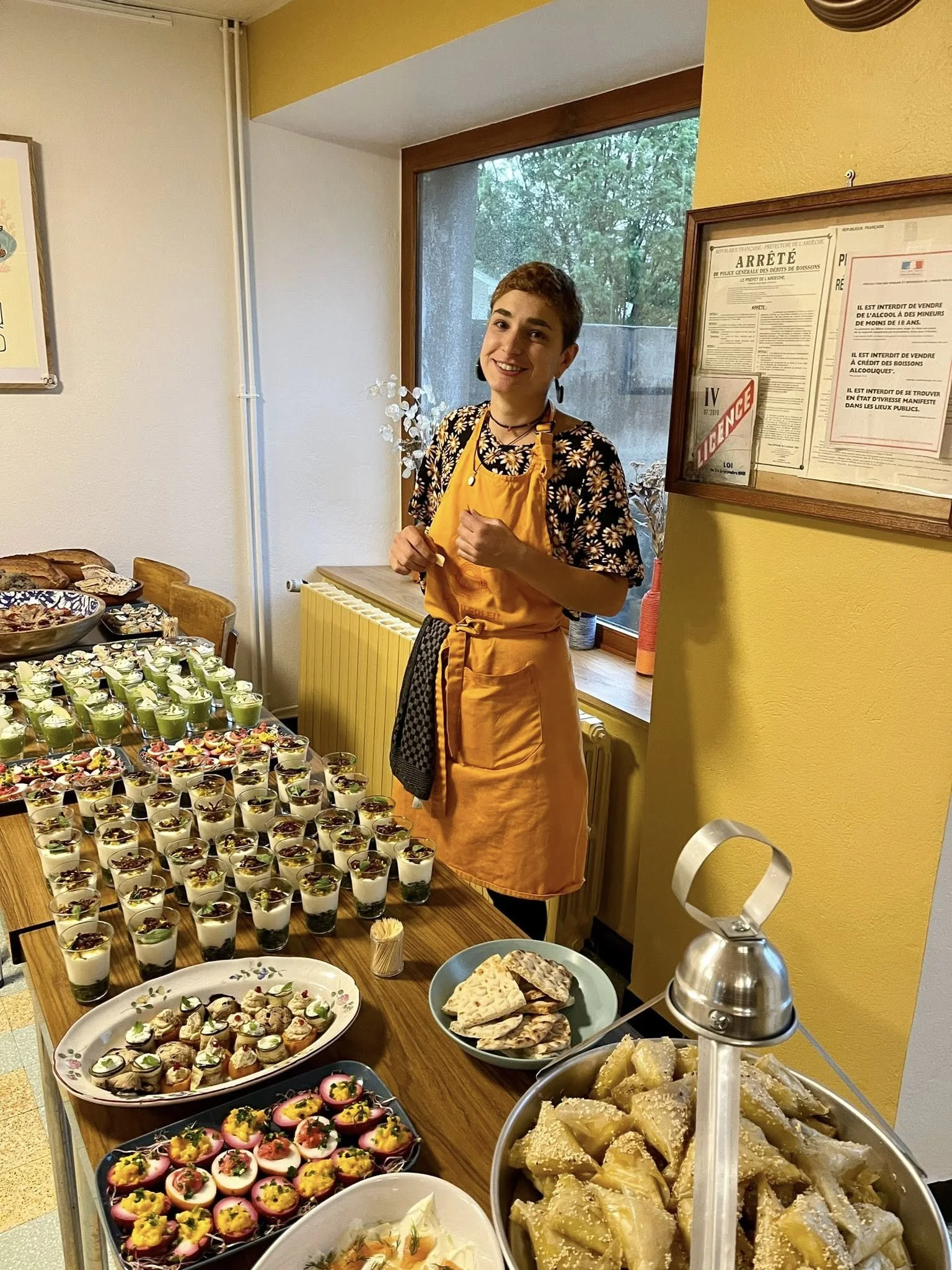 Une jeune femme souriante portant un tablier jaune se tient derrière une table remplie de desserts dans une pièce lumineuse avec une grande fenêtre. La table présente une variété de verrines, cupcakes, biscuits, et autres douceurs, accompagnés de cou