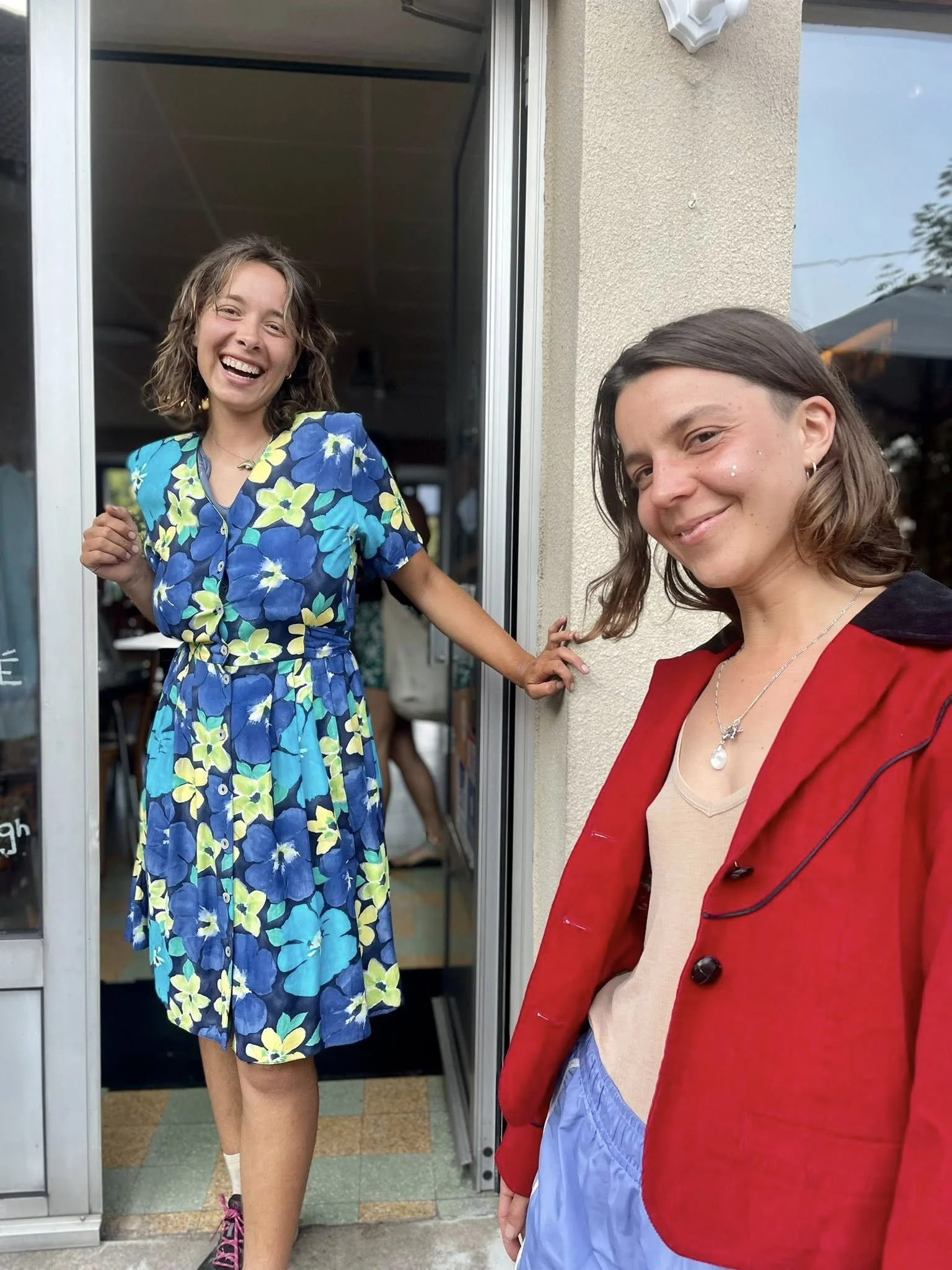 Deux jeunes femmes souriantes près d'une porte d'entrée, une portant une robe à fleurs bleues et vertes, l'autre une veste rouge. En arrière-plan, à l'intérieur, on voit des personnes et une ambiance décontractée.