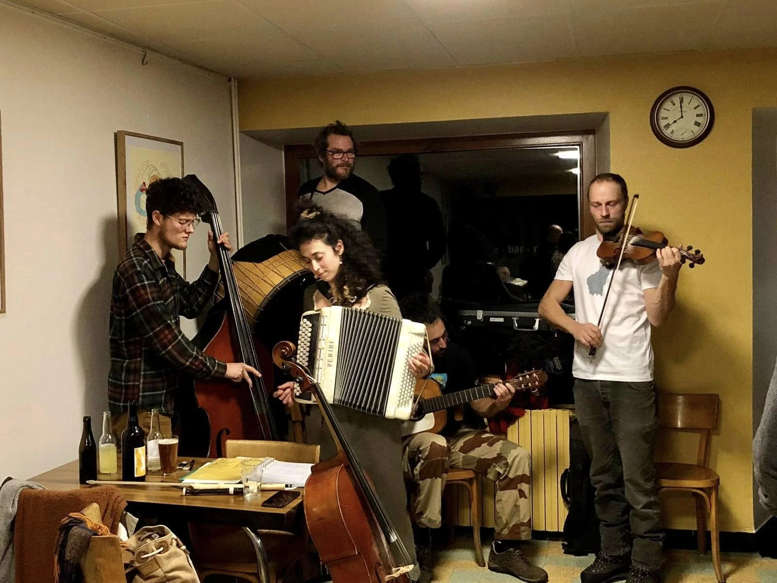 Un groupe de musiciens jouant ensemble dans une pièce, avec un piano en arrière-plan et des boissons sur une table.