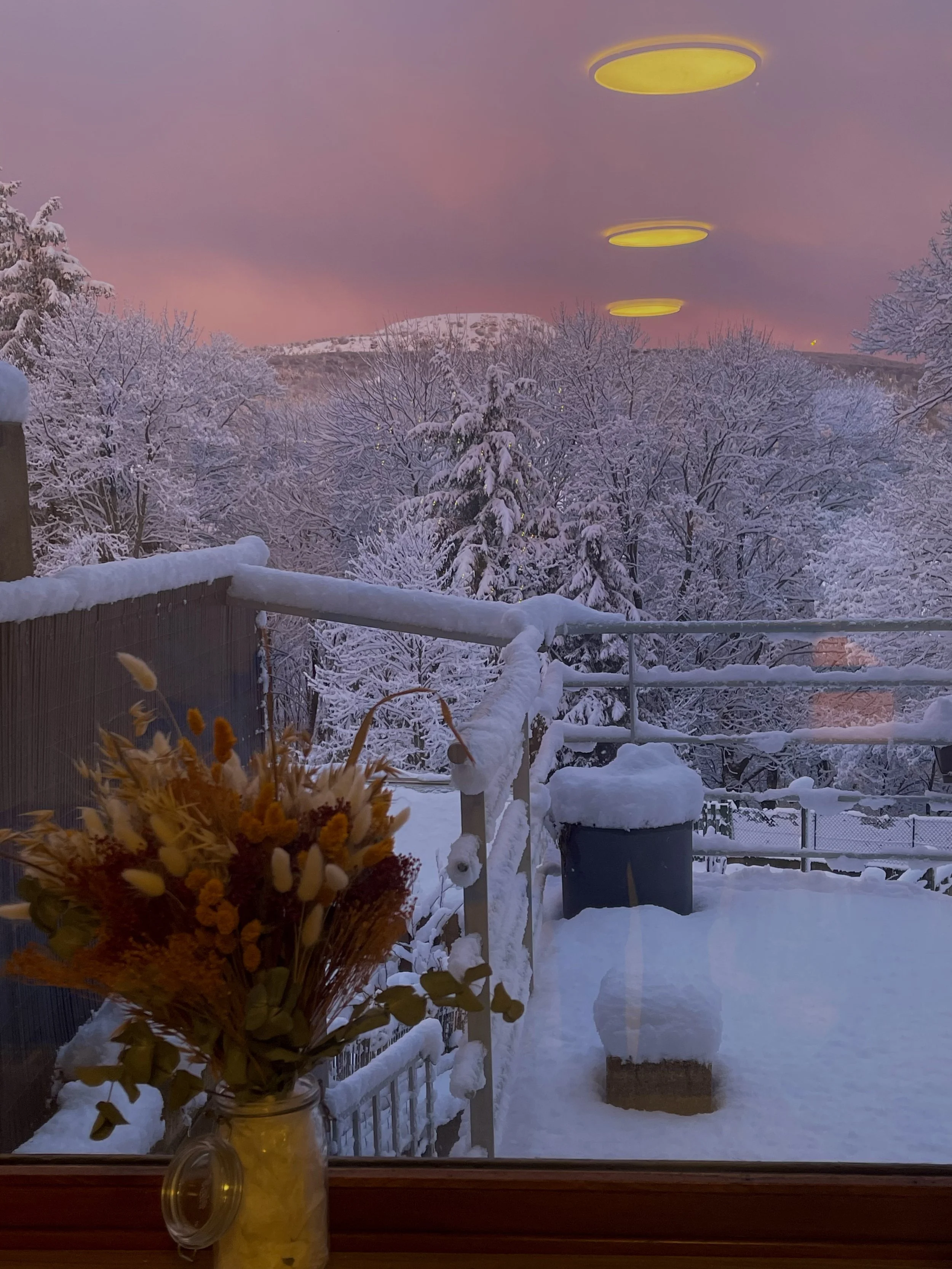 Vue d'une terrasse enneigée avec des arbres couverts de neige, un ciel rose au coucher du soleil, un vase de fleurs à l'avant, et trois plafonniers jaunes en reflet sur la vitre.
