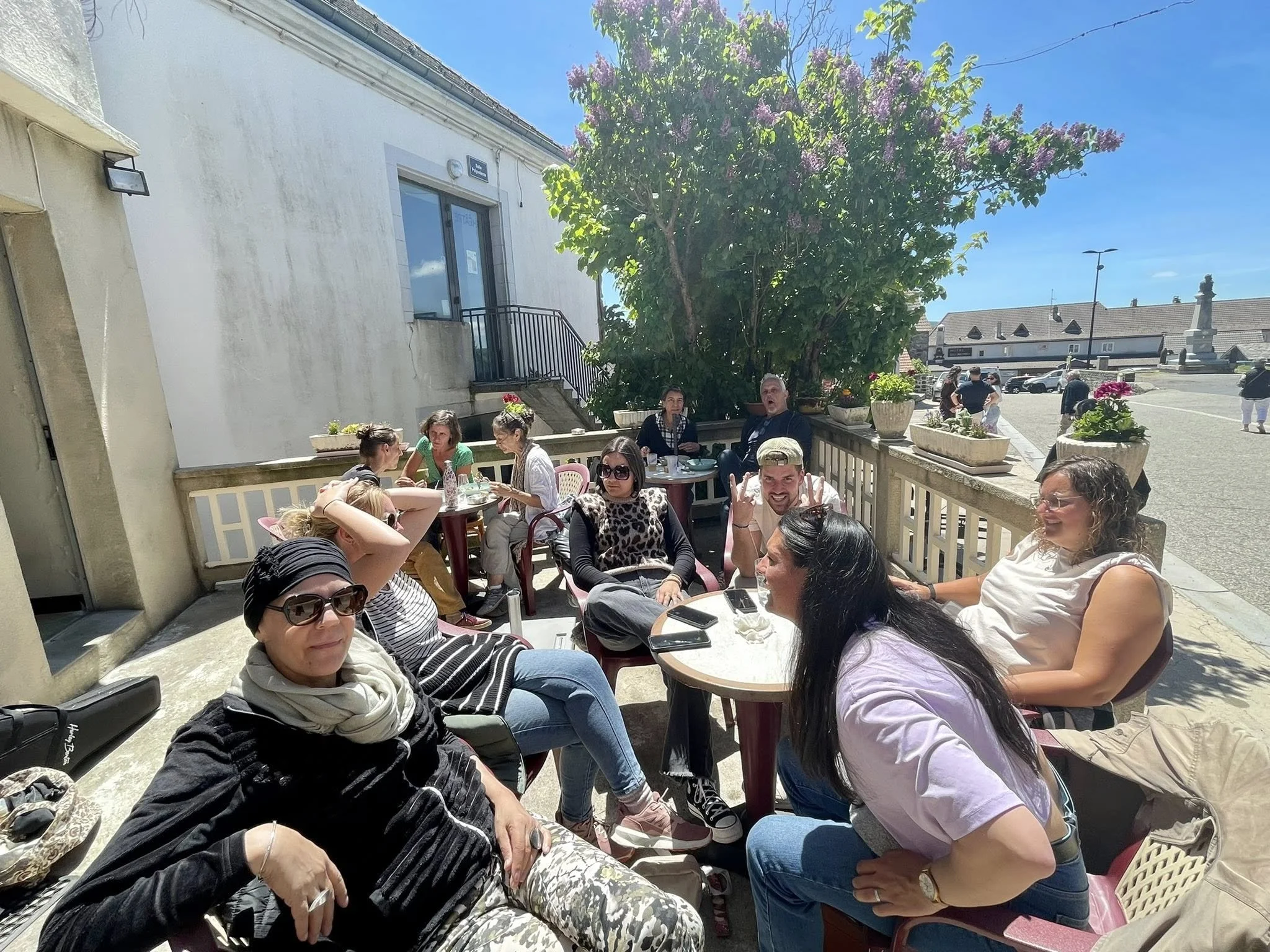 Groupe de personnes assises à une table en extérieur, en train de discuter et de profiter du soleil, avec un arbre fleuri en arrière-plan.