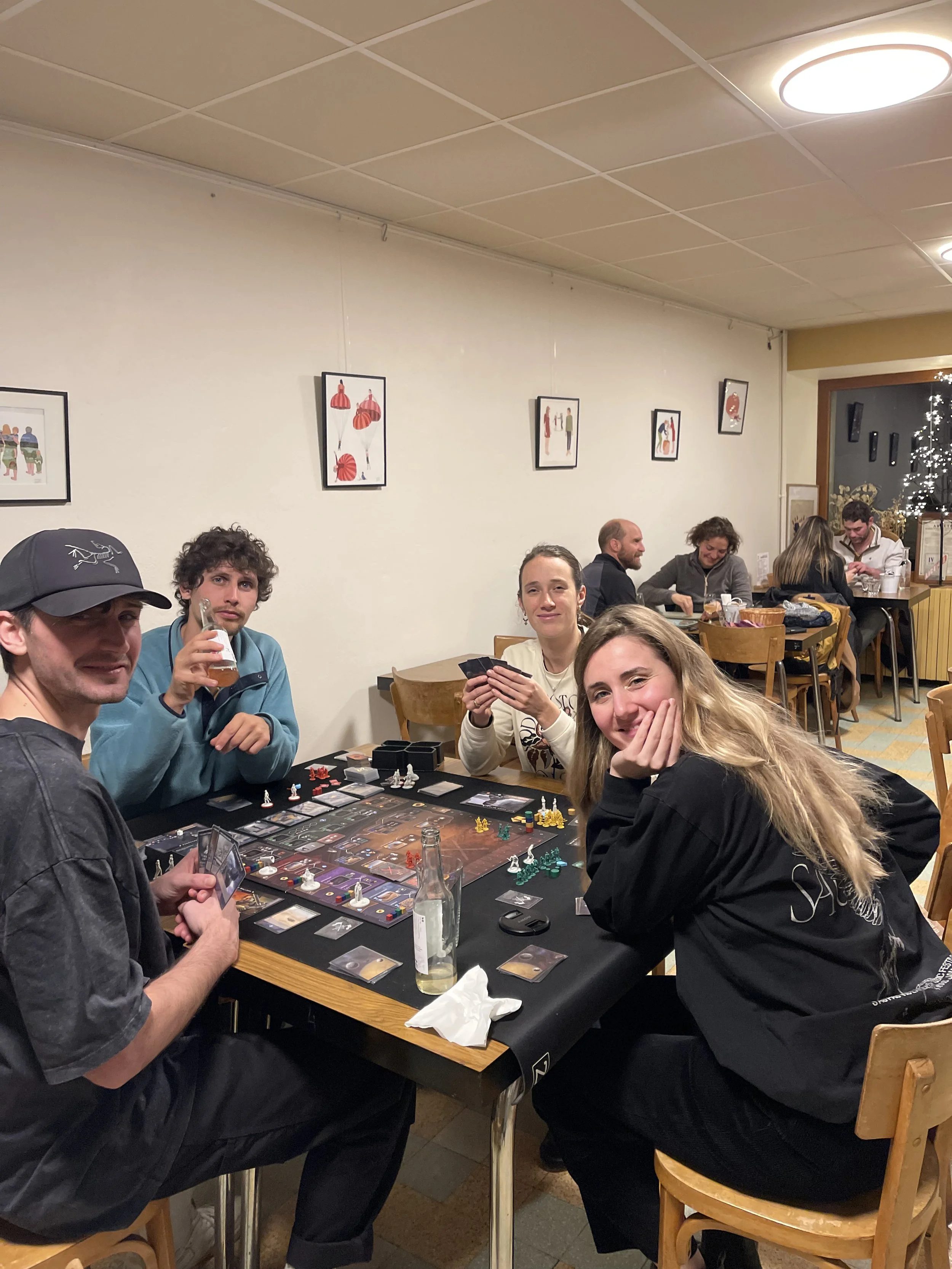 Groupe de quatre personnes jouant à un jeu de société dans un restaurant ou café. La table est remplie de pièces de jeu et de cartes, avec une bouteille et des verres. Quatre autres personnes sont visibles à l'arrière, discutant et profitant de leur 