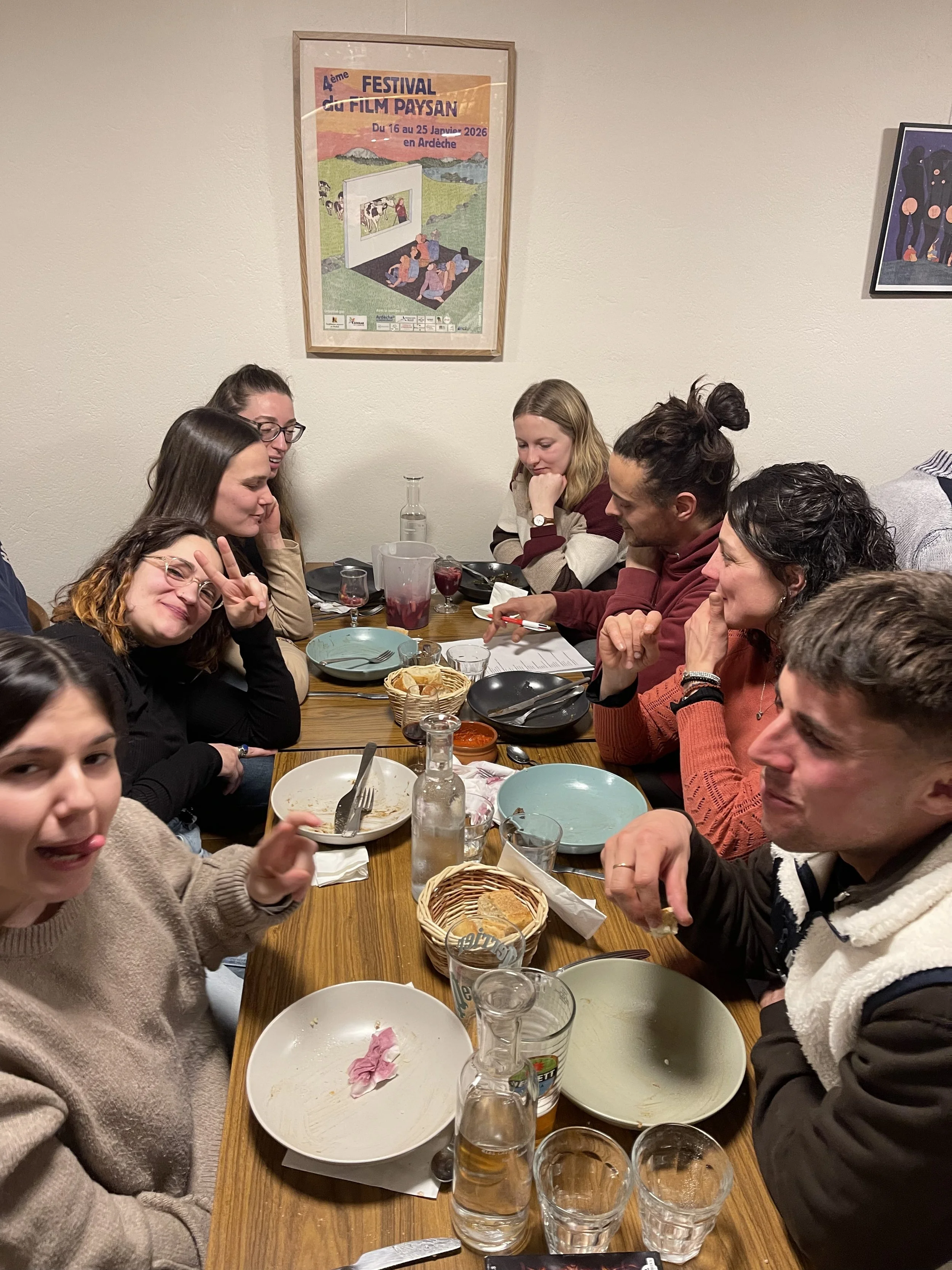 Groupe de personnes assises autour d'une table, partageant un repas, avec des assiettes vides, des verres, des bouteilles et des snacks, dans une salle avec des posters au mur.