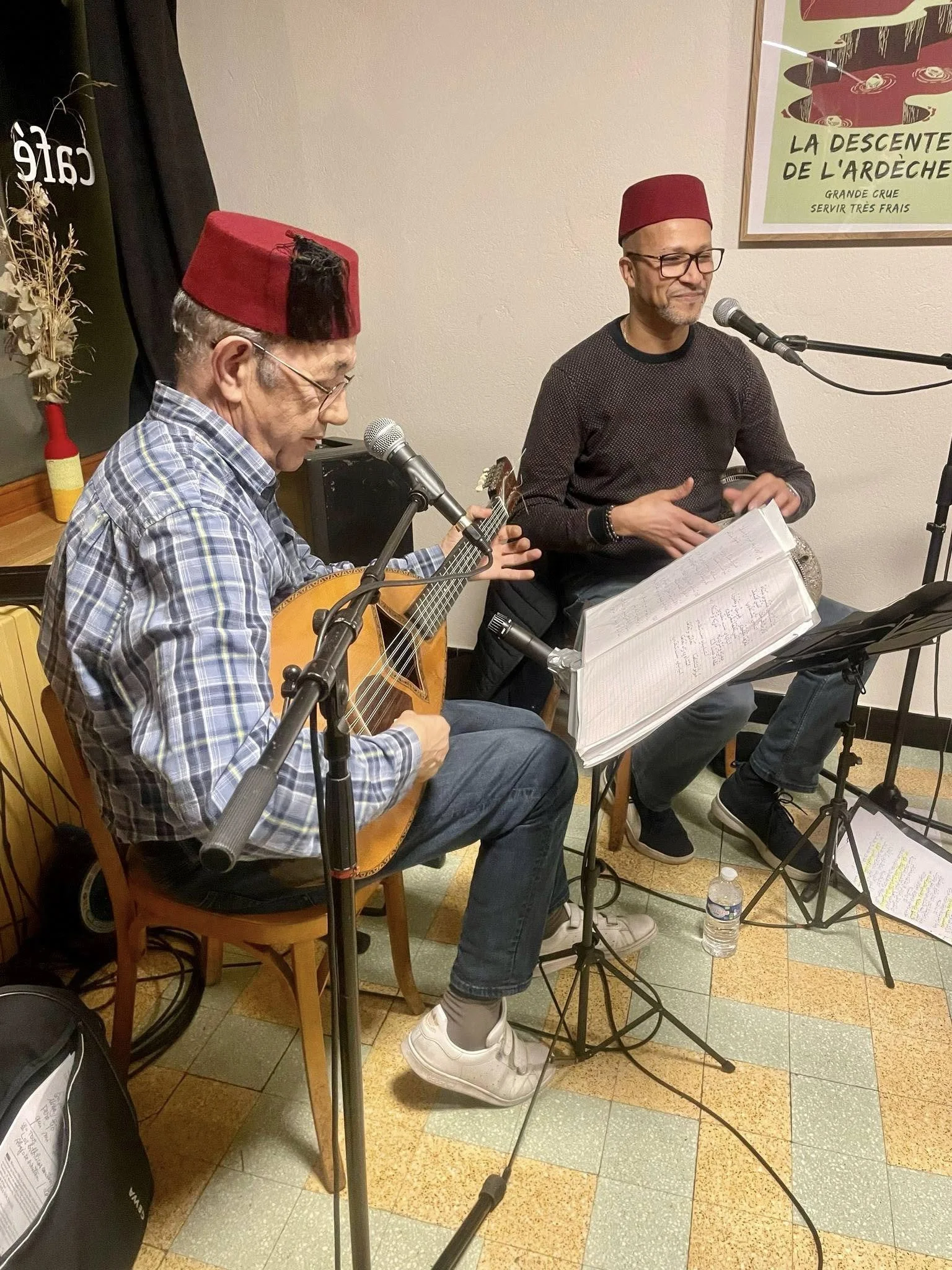 Deux hommes chantent et jouent de la guitare dans une salle, portant tous deux une fez rouge. L'un des hommes joue de la guitare, tandis que l'autre chante derrière un micro. Il y a un cadre avec une affiche en arrière-plan.