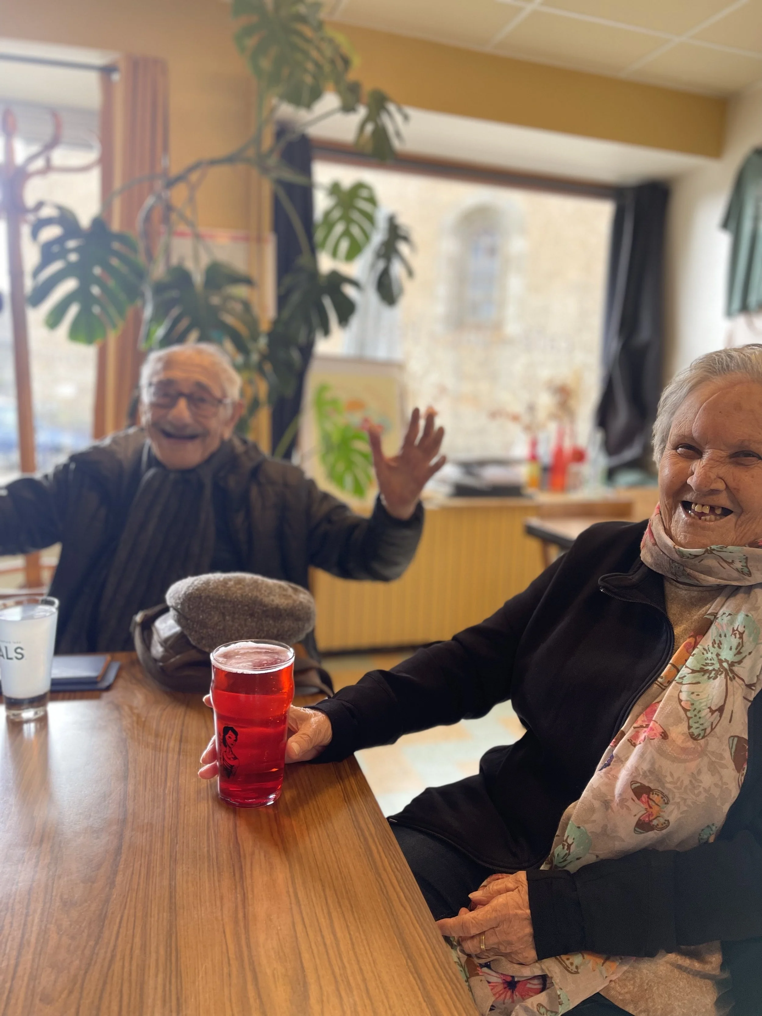 Deux personnes âgées souriantes assises à une table en bois, une d'elles tient un verre de boisson rouge. Elles semblent ravies et en train de partager un moment joyeux.