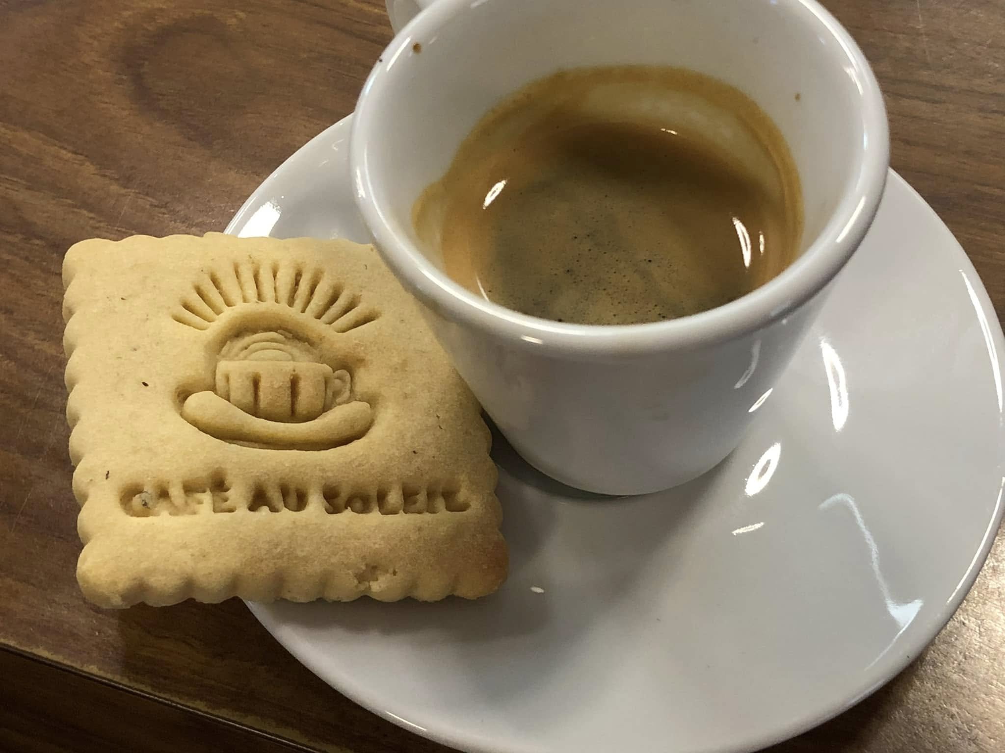 Une tasse de café espresso vide sur une assiette blanche avec un biscuit rectangulaire en forme de logo Château d'Olères, avec une image d'un visage souriant et le texte "CHATEAU OLÈRES" en dessous.
