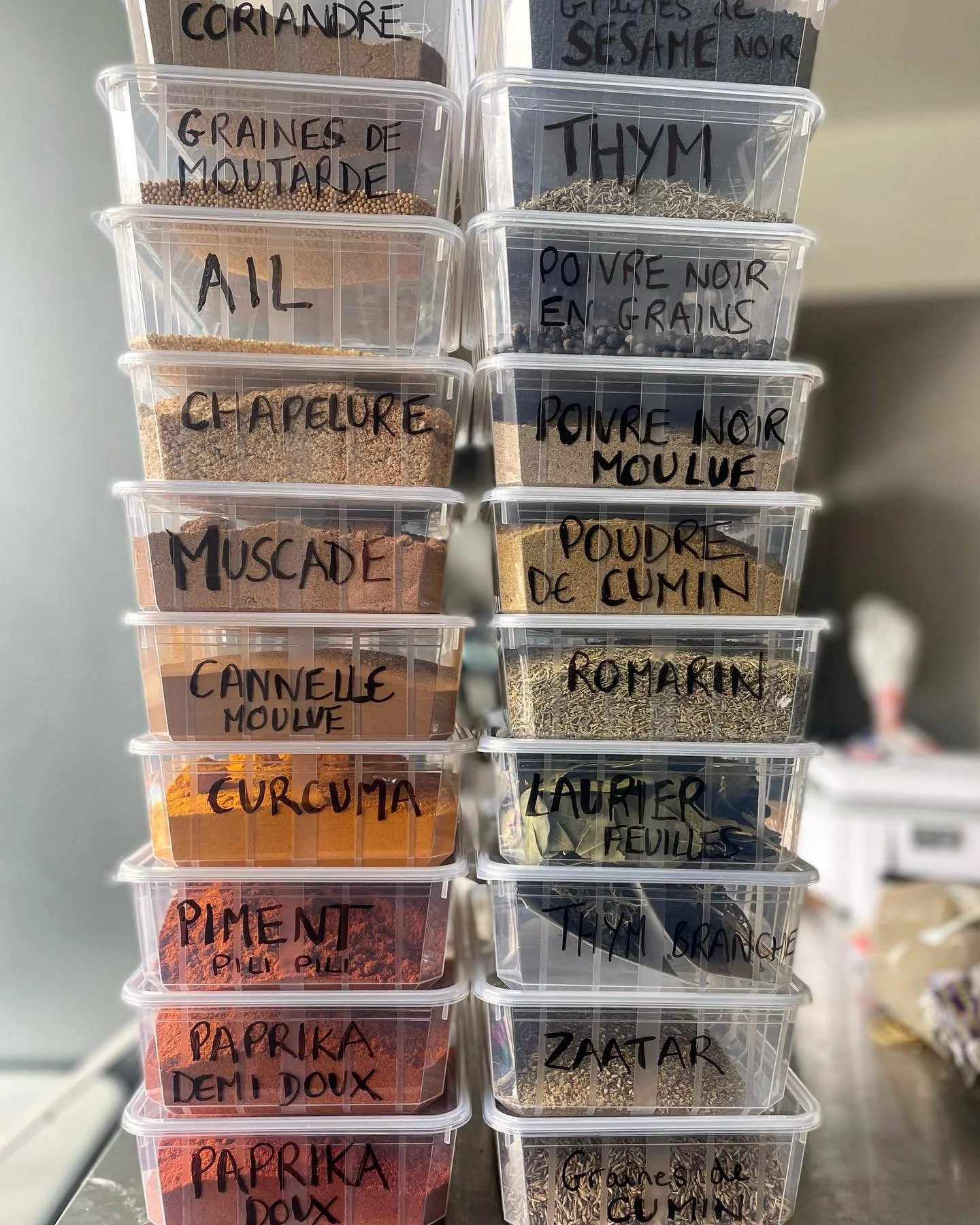 Étagero avec des boîtes en plastique contenant des épices séchées et des herbes, avec des étiquettes écrites à la main indiquant leur contenu en français.