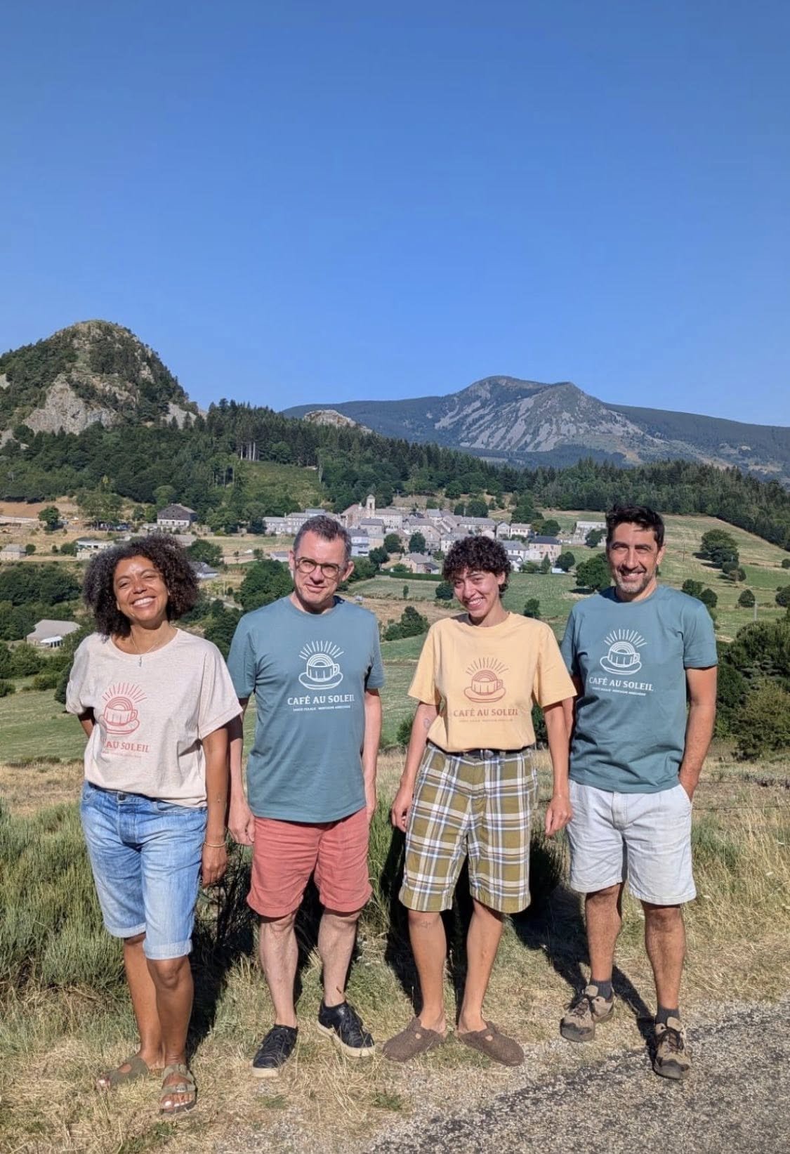 Groupe de quatre personnes debout dans un champ avec une montagne en arrière-plan, tous portant des t-shirts colorés avec le logo 'Café au Soleil'