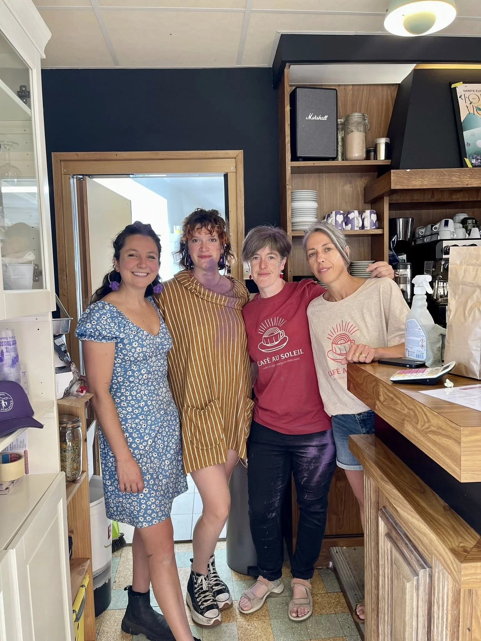 Quatre femmes souriantes debout dans un café, posant pour la photo. L'une porte une robe bleue à motifs floraux, une autre une robe à rayures marron et jaunes, une troisième une t-shirt rouge avec le texte « Café au Soleil », et la dernière un t-shir