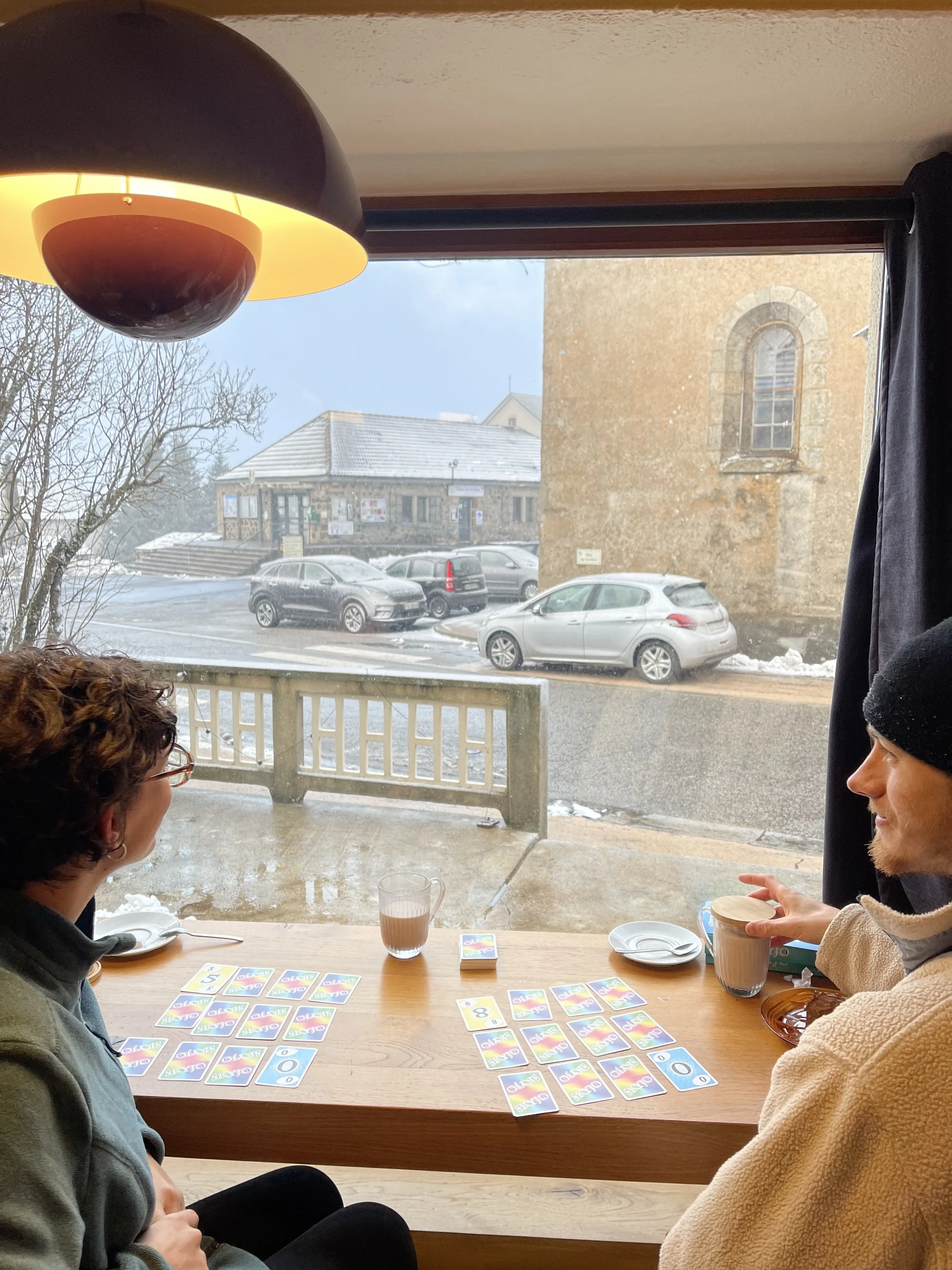 Deux personnes jouent à un jeu de cartes avec des cartes colorées sur une table en bois dans un café, avec des boissons chaudes. La fenêtre derrière montre une rue enneigée avec des voitures stationnées et un bâtiment en pierre.