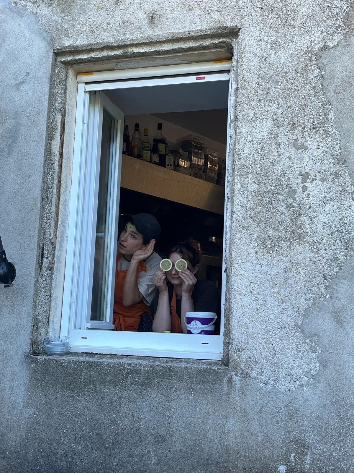 Deux personnes regardant par une fenêtre, portant des rondelles de citron sur leurs yeux, dans une cuisine.