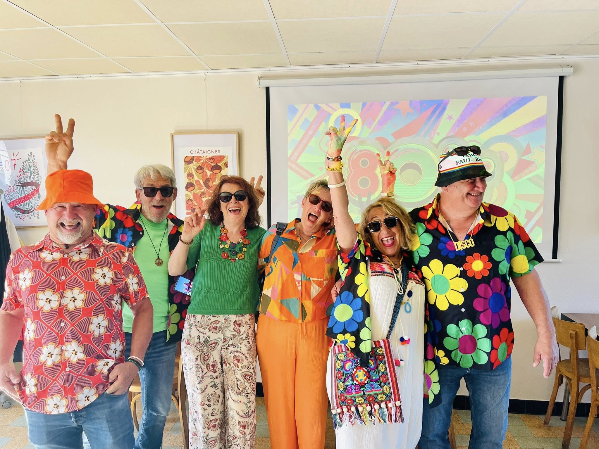 Groupe de six personnes déguisées en style coloré et rétro lors d'une fête ou d'une célébration, posant avec des expressions joyeuses et gesticulant, dans une salle avec un écran de projection et des affiches à l'arrière.