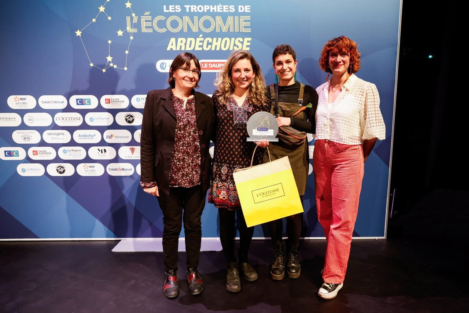 Groupe de cinq femmes posant lors d'une cérémonie de remise de trophée pour l'économie en Ardèche, avec un trophée et un sac cadeau de la marque L'Occitane, devant une bannière avec des logos de sponsors.