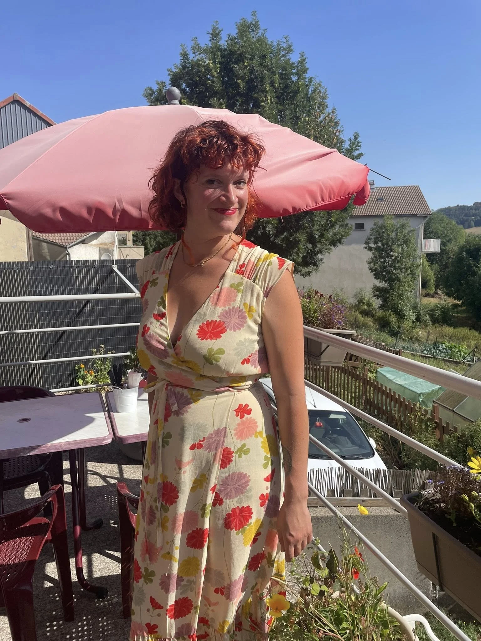 Une femme en robe à fleurs pose sur une terrasse ensoleillée avec un parasol rose, des plantes en pots, une voiture blanche et des maisons en arrière-plan.