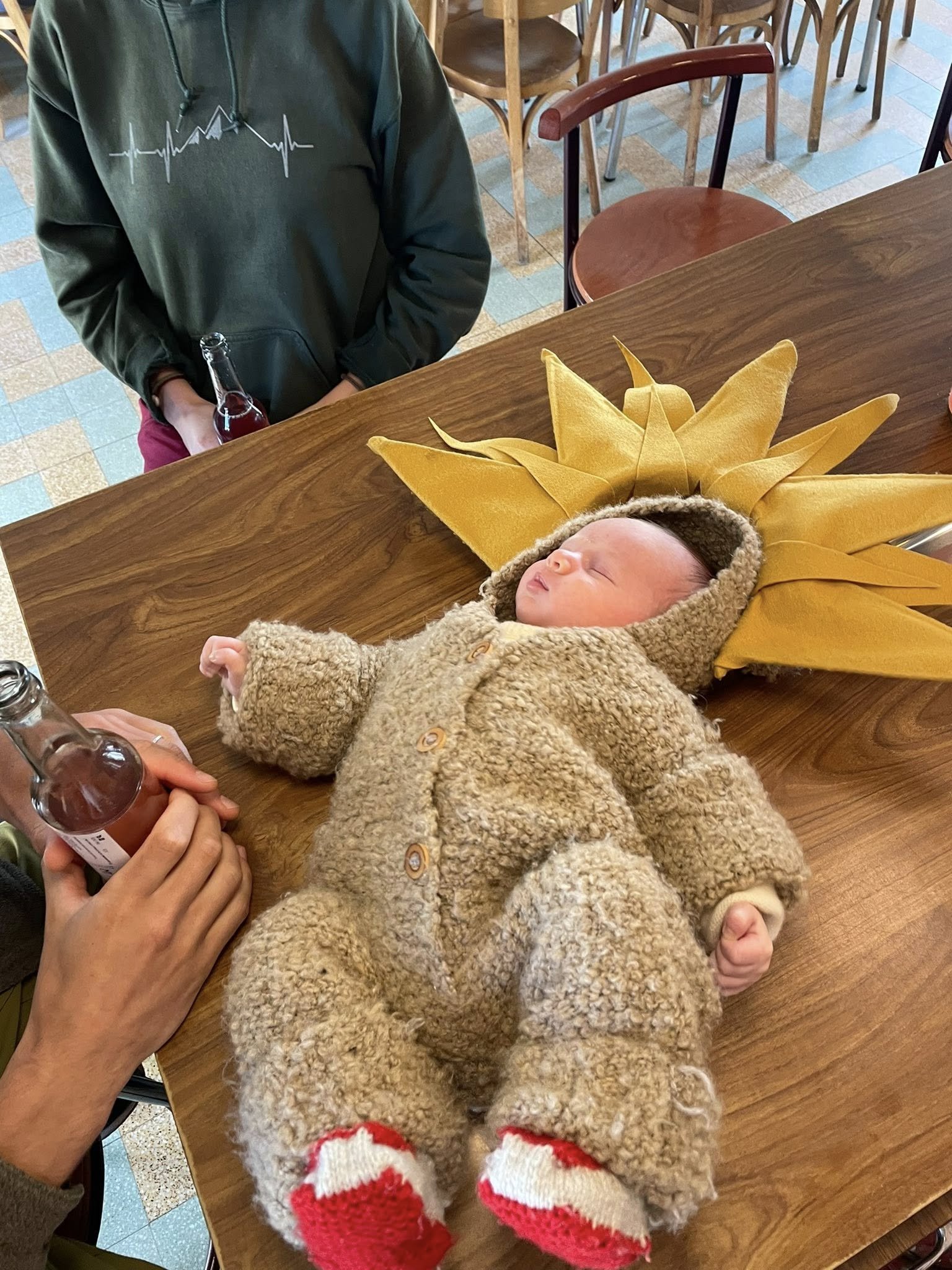 Un bébé endormi vêtu d'un costume en laine beige avec un bonnet et des chaussettes rouges et blanches, allongé sur une table en bois, avec un halo en forme de soleil jaune derrière lui.