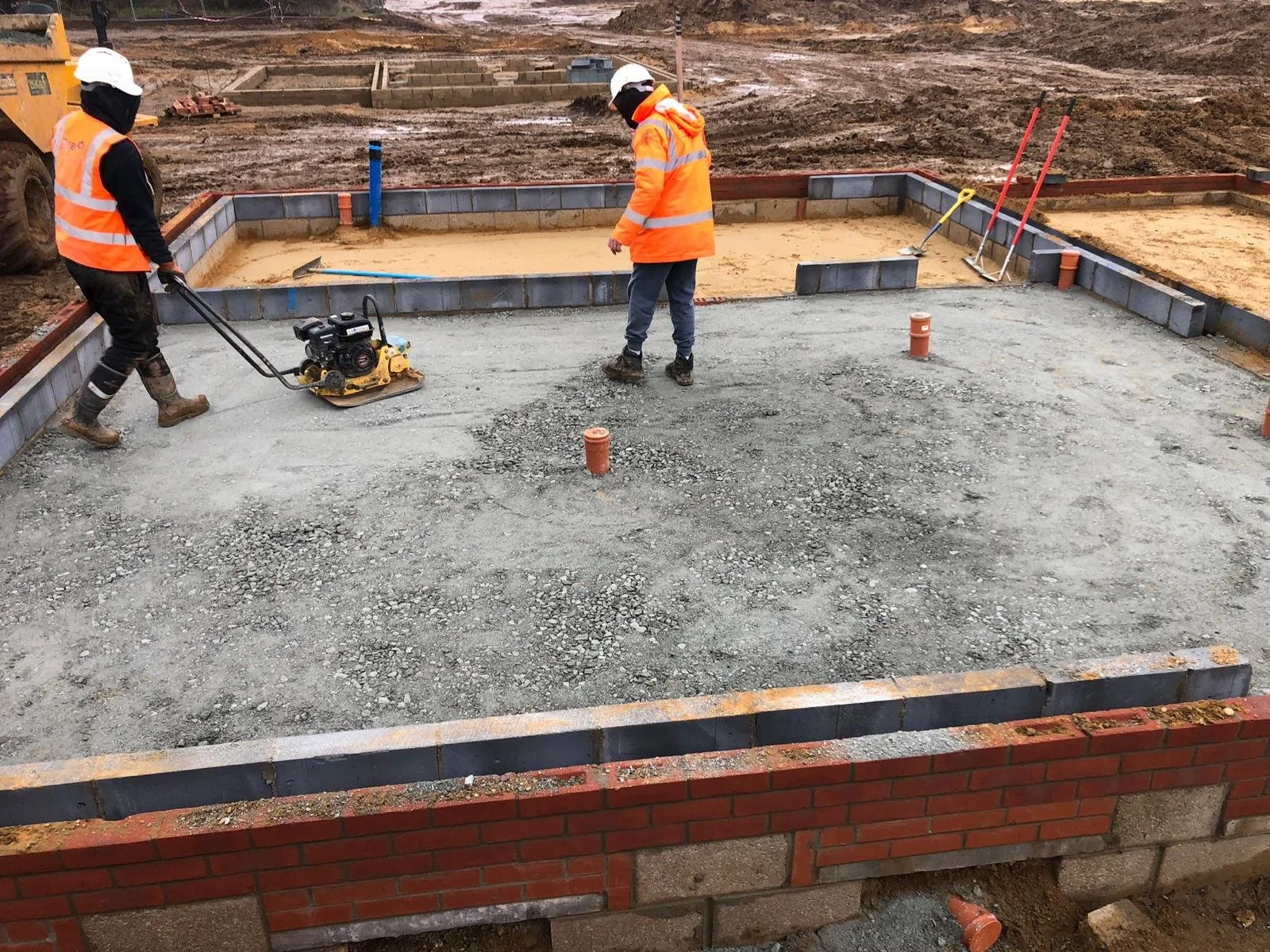 Construction workers in orange safety vests and helmets working on a building foundation with a concrete-like surface, surrounded by brick and cinder block walls, with construction tools and equipment on site.