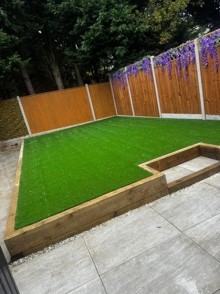 A backyard with artificial grass, a wooden raised garden bed, a wooden fence with purple flowering vines, and surrounded by trees.