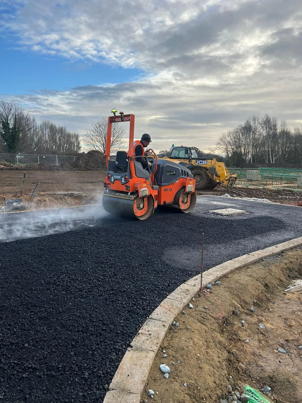 Construction worker operating a small compactor on a newly paved road with a JCB construction vehicle in the background under a cloudy sky.
