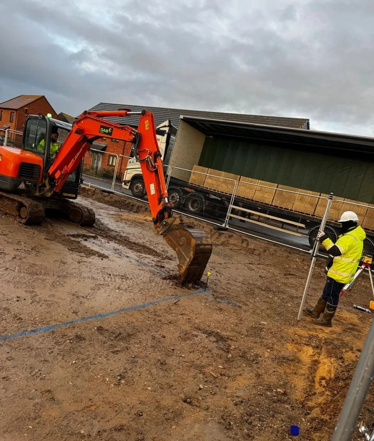 Construction site with a small orange excavator digging in the dirt and a worker in a yellow high-visibility jacket and white helmet taking measurements nearby.