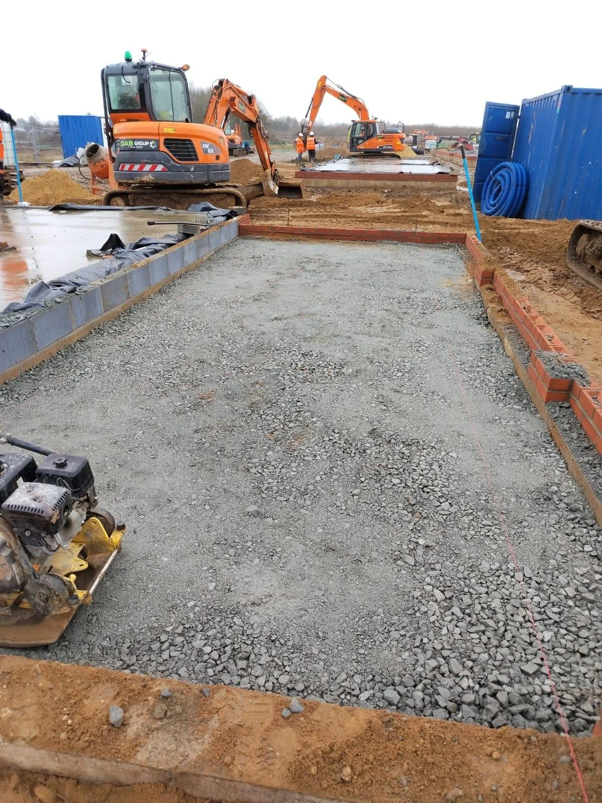 Construction site with excavators working, gravel being leveled, and brick borders being built for a foundation.