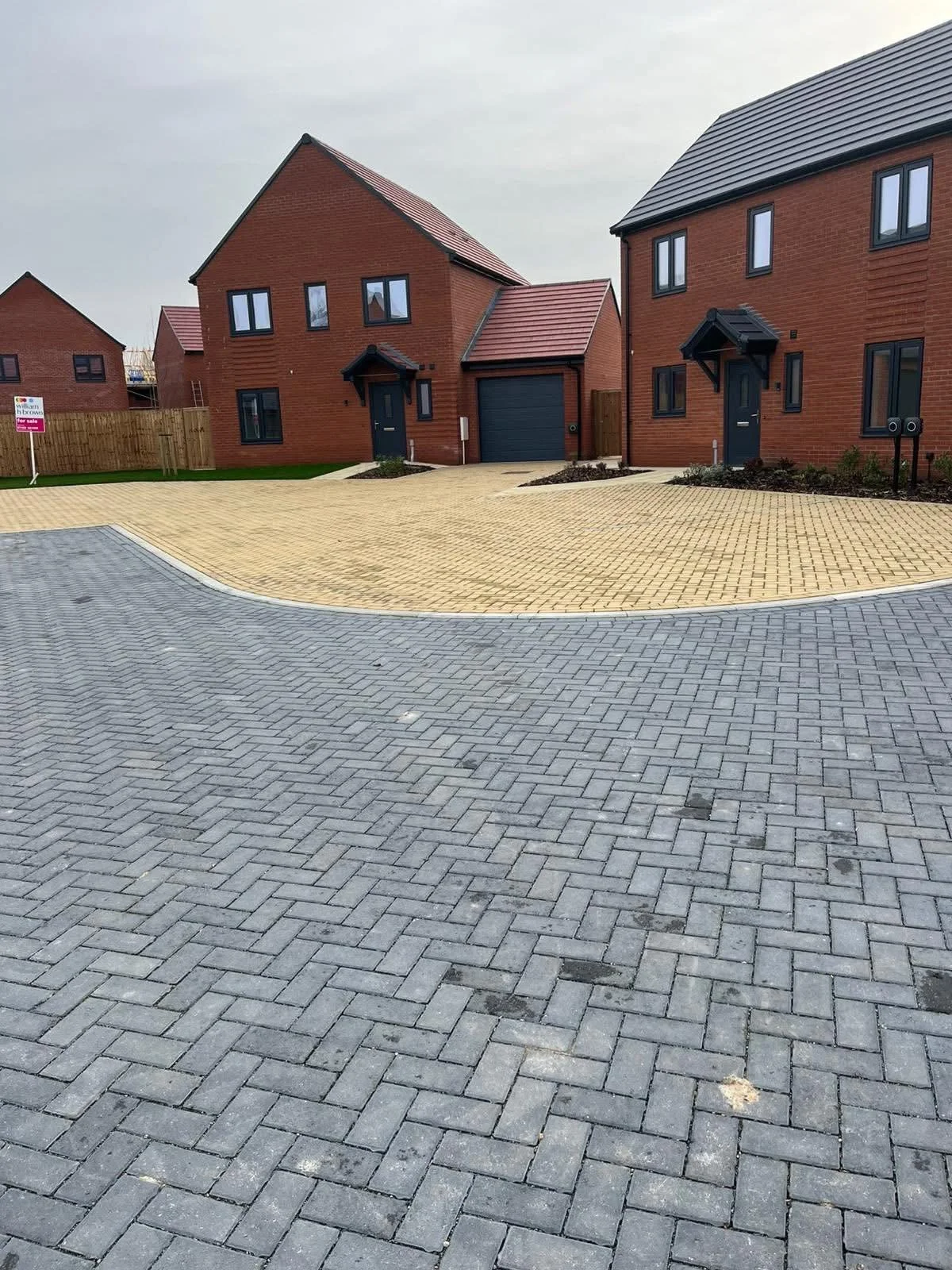 Newly built two-story brick houses with dark roofs, black doors, and a driveway with interlocking pavers, under a cloudy sky.