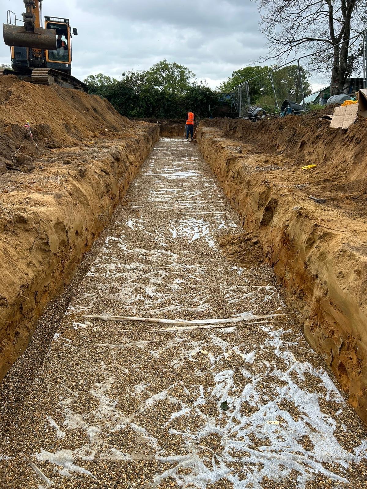 Construction site with excavated trench and a worker in orange vest, excavator on the side, dirt and gravel, overcast sky.