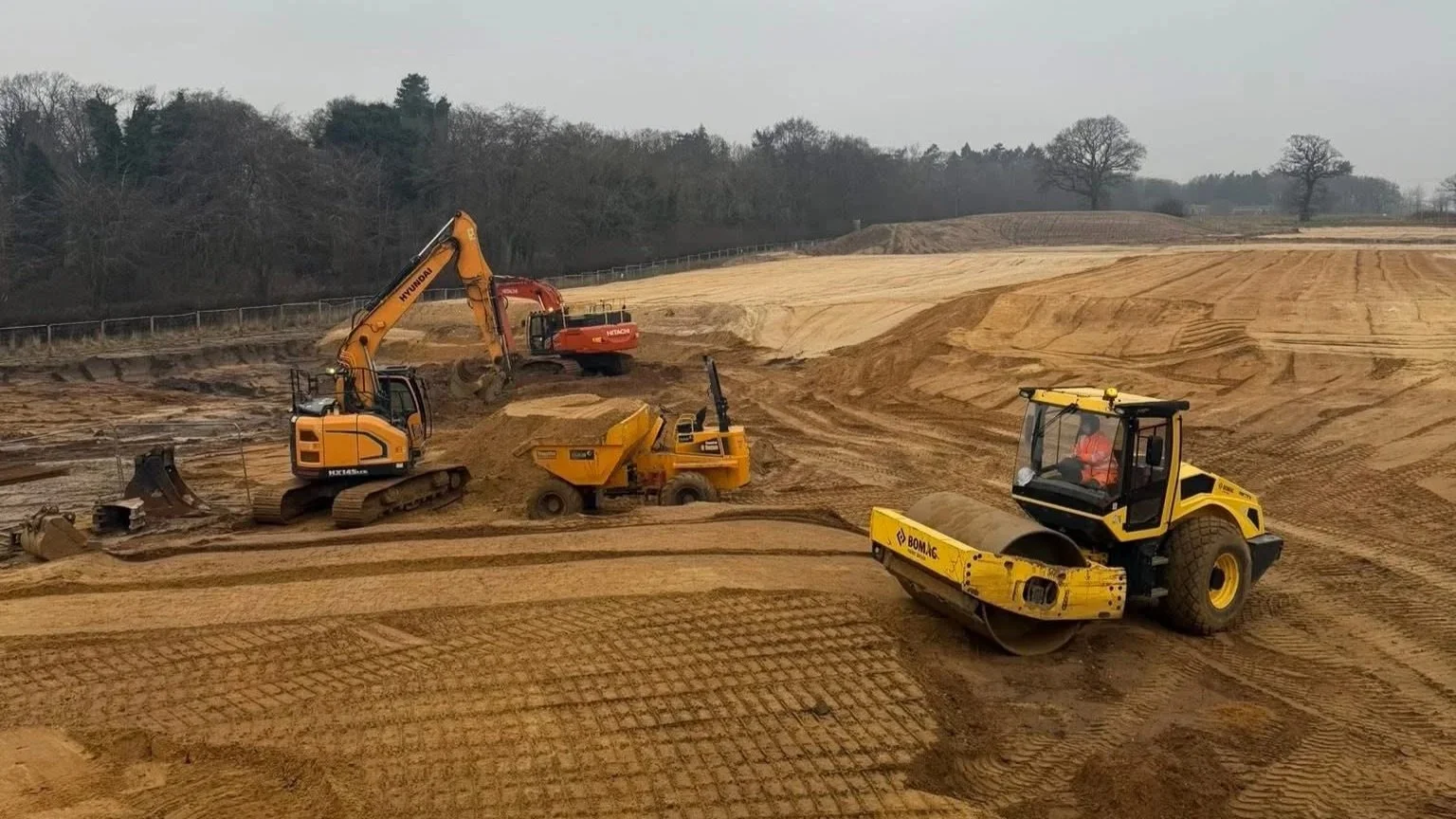 Construction site with multiple heavy machinery, including an excavator, dumper, and roller, moving and compacting dirt on a sloped terrain.