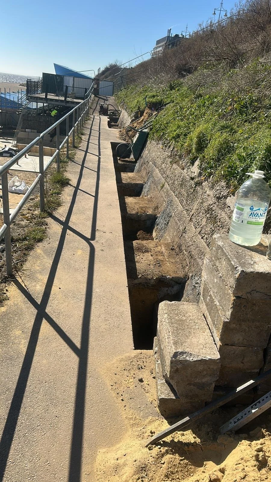Sidewalk with trench and construction tools along a cliff face with greenery, a water bottle placed on the concrete, and a railing on the left side, under clear sunny sky.
