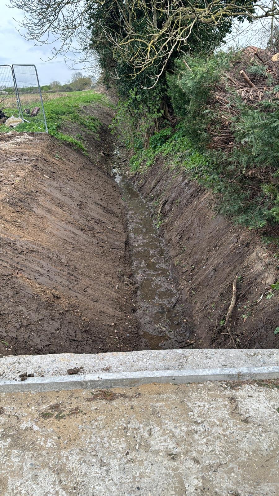 A narrow ditch with a newly installed culvert in wet, muddy soil, surrounded by greenery and trees, with a concrete barrier at the foreground.