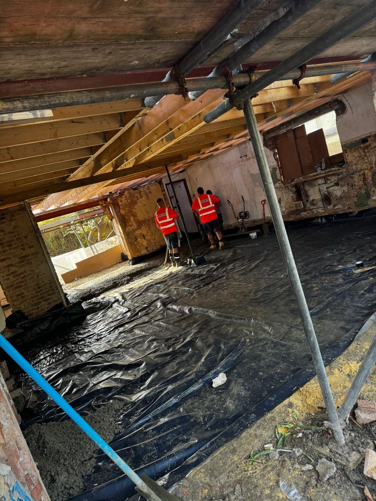 Three workers in red safety vests and black shorts working inside a building under construction, with exposed brick walls, wooden ceiling framing, and a black plastic ground covering.