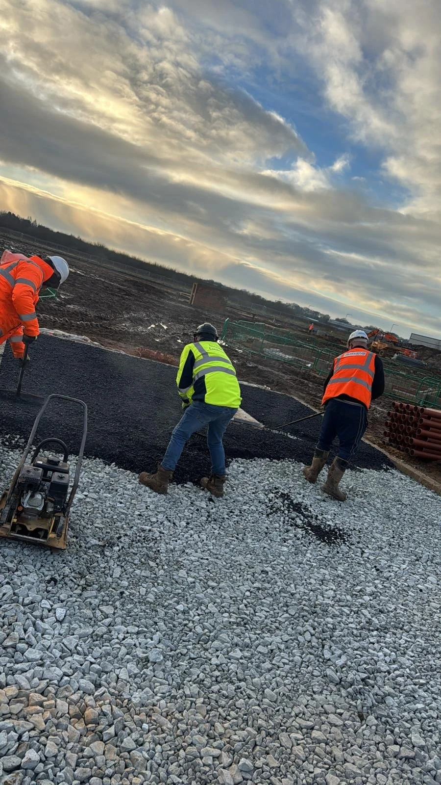 Construction workers paving a road with asphalt and gravel, wearing safety vests and helmets, under a partly cloudy sky.